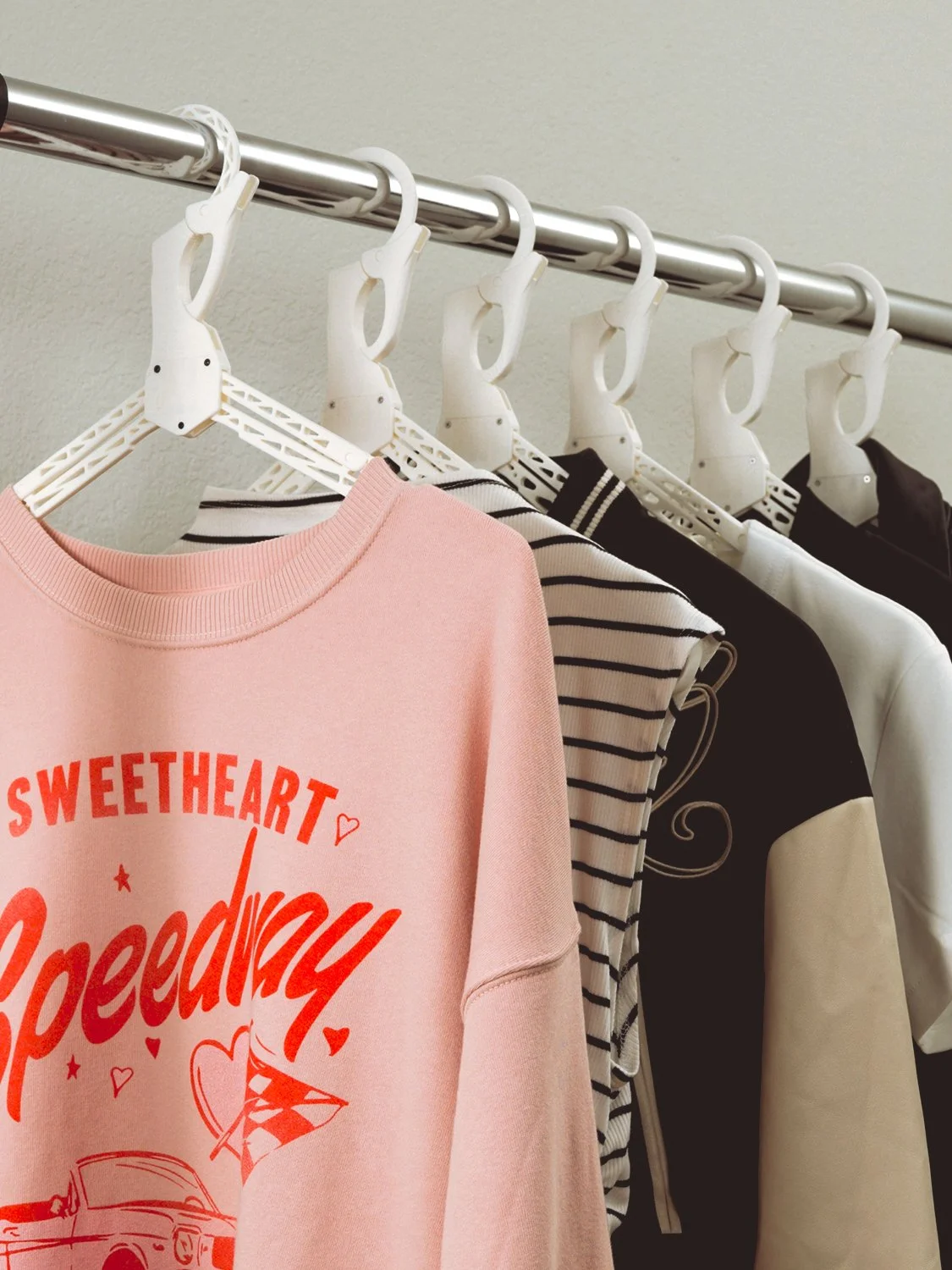 A pink sweatshirt with red text and graphics hanging on a white plastic hanger, alongside striped and solid color shirts on a clothing rack.