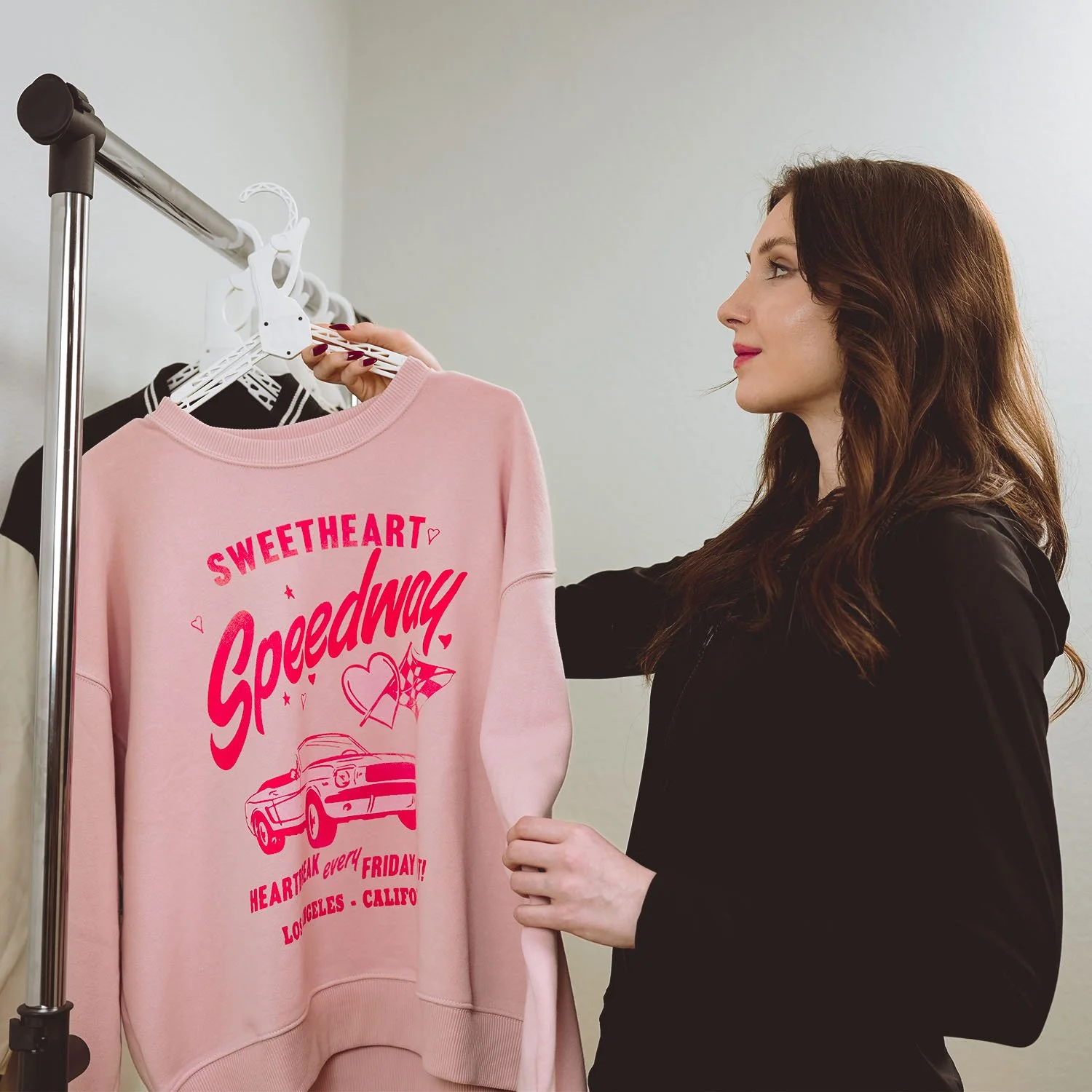 Woman with long brown hair, wearing a black jacket, holds a pink sweatshirt on a hanger. The sweatshirt has red text and graphics related to a race event, including a car and the words 'Sweetheart Speedway' and 'Heart Trak every Friday! Los Angeles - Califo'.