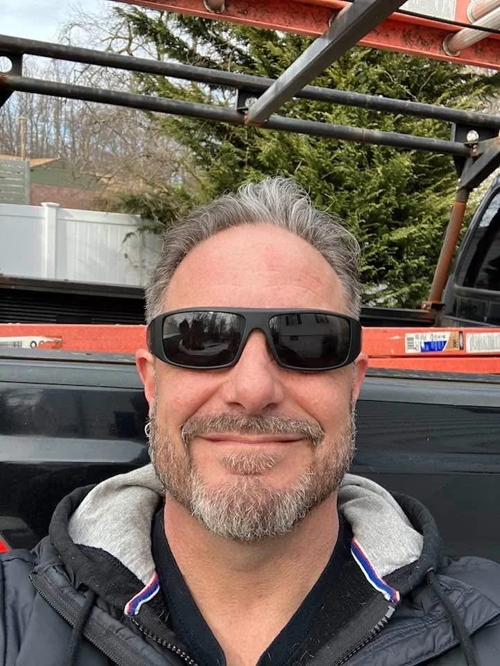 A man with gray hair and a beard smiling at the camera, wearing sunglasses and a black jacket, standing in front of a black vehicle with a ladder on top in an outdoor setting.