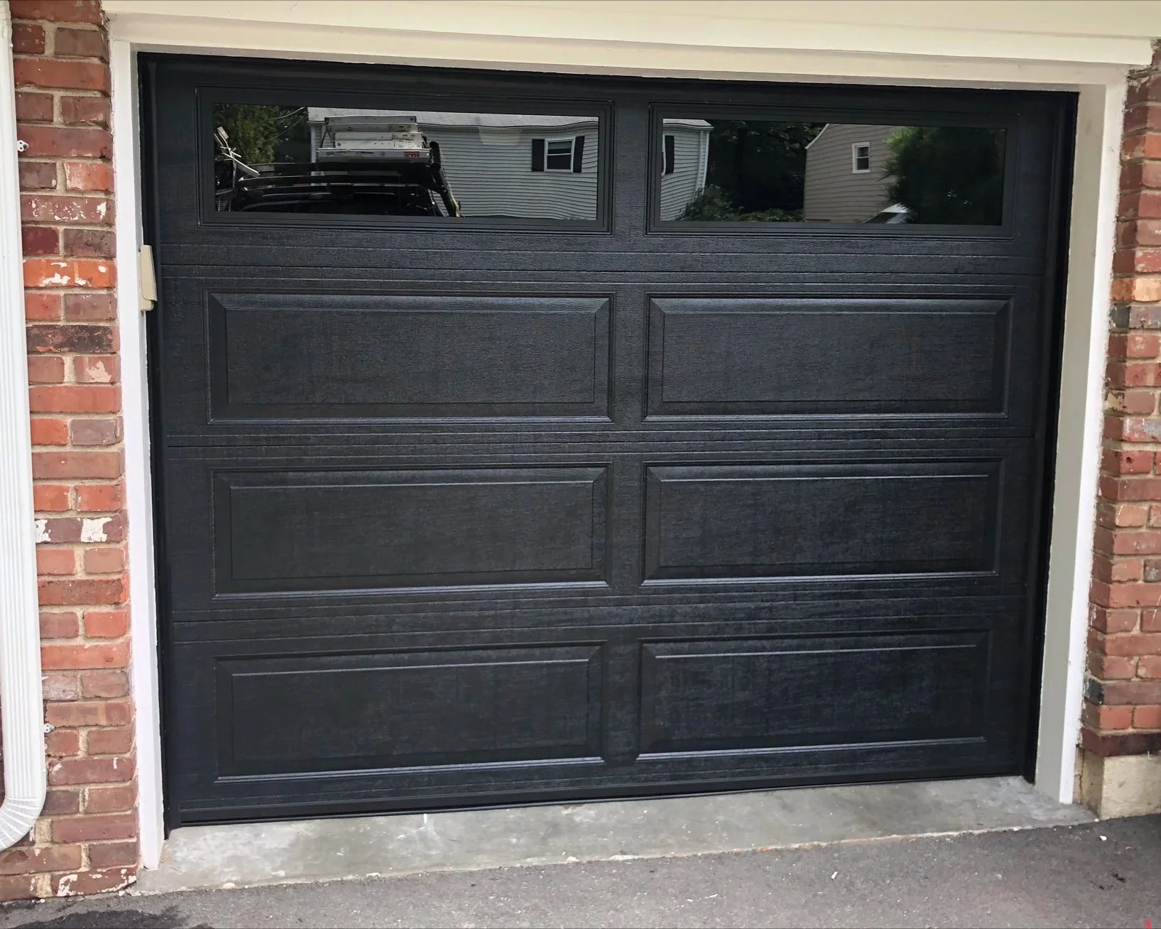 Black garage door with four rectangular panels and a row of small windows at the top, set in a brick house with white trim.