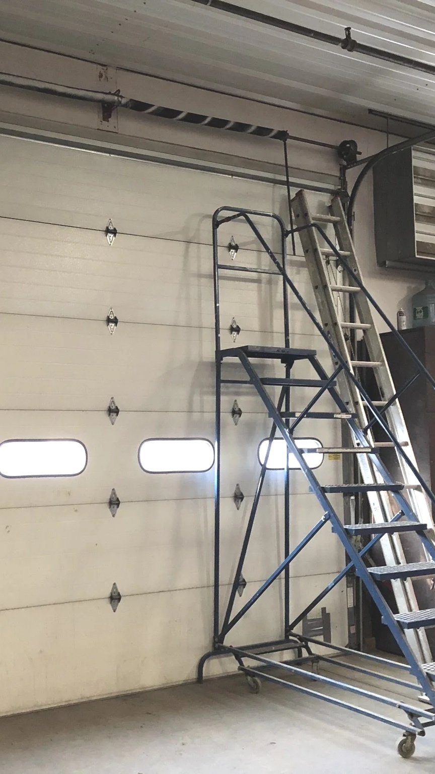 A garage with a closed roll-up door, a rolling scaffold, a ladder, and a garage heater mounted on the wall.