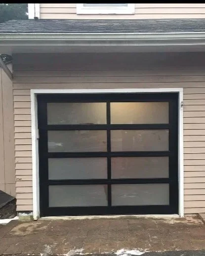 Black-framed garage door with six frosted glass panels, beige house siding, and a concrete driveway.