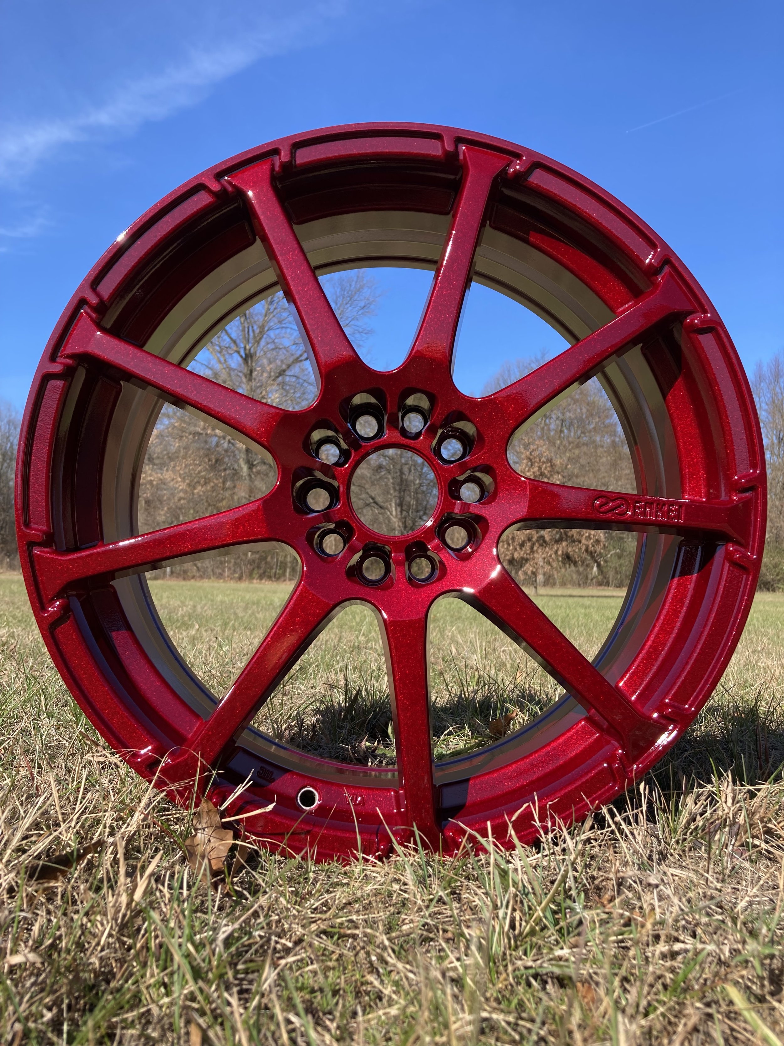 Red alloy wheel rim on grass field outdoors with blue sky and trees in background.