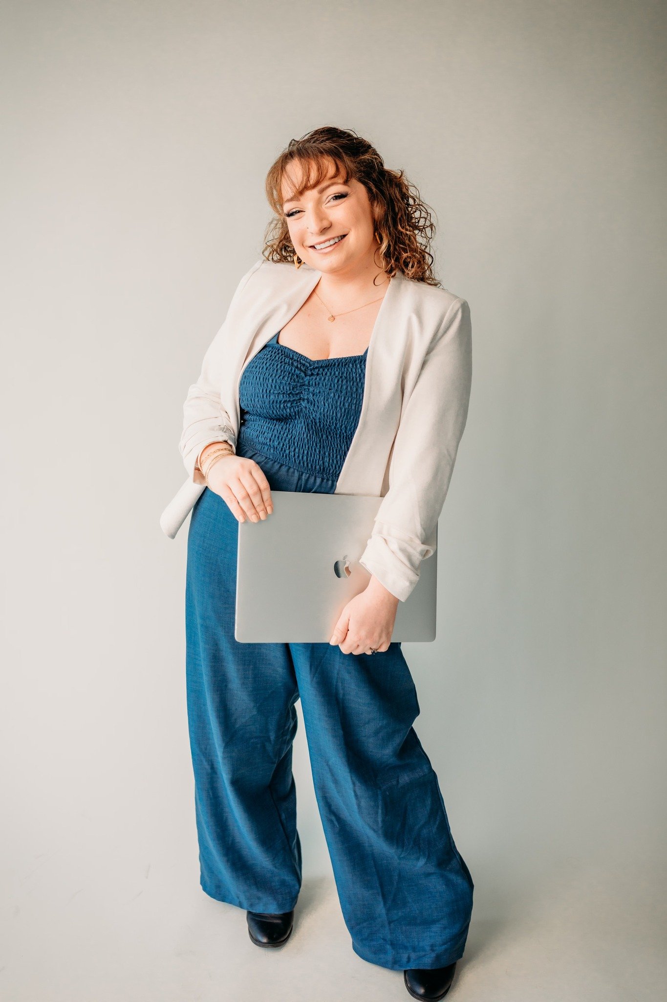 Woman smiling, holding silver laptop, dressed in a cream blazer, blue smocked top, and wide-leg blue pants, standing against a plain light background.