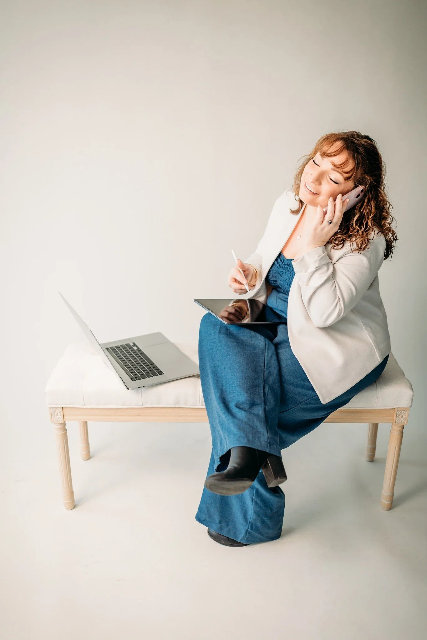 A woman sitting on a bench, smiling and talking on her cell phone, with a tablet and a laptop nearby.