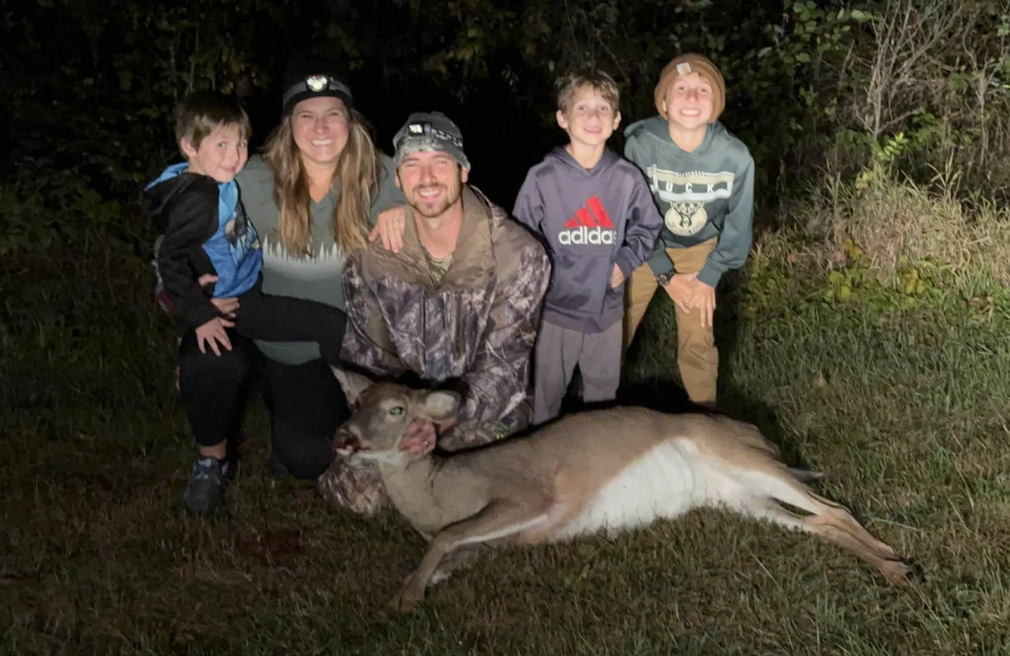 A group of six people, including children and adults, posing outdoors at night around a deceased deer. They are smiling and kneeling or standing on grass with dark foliage in the background.