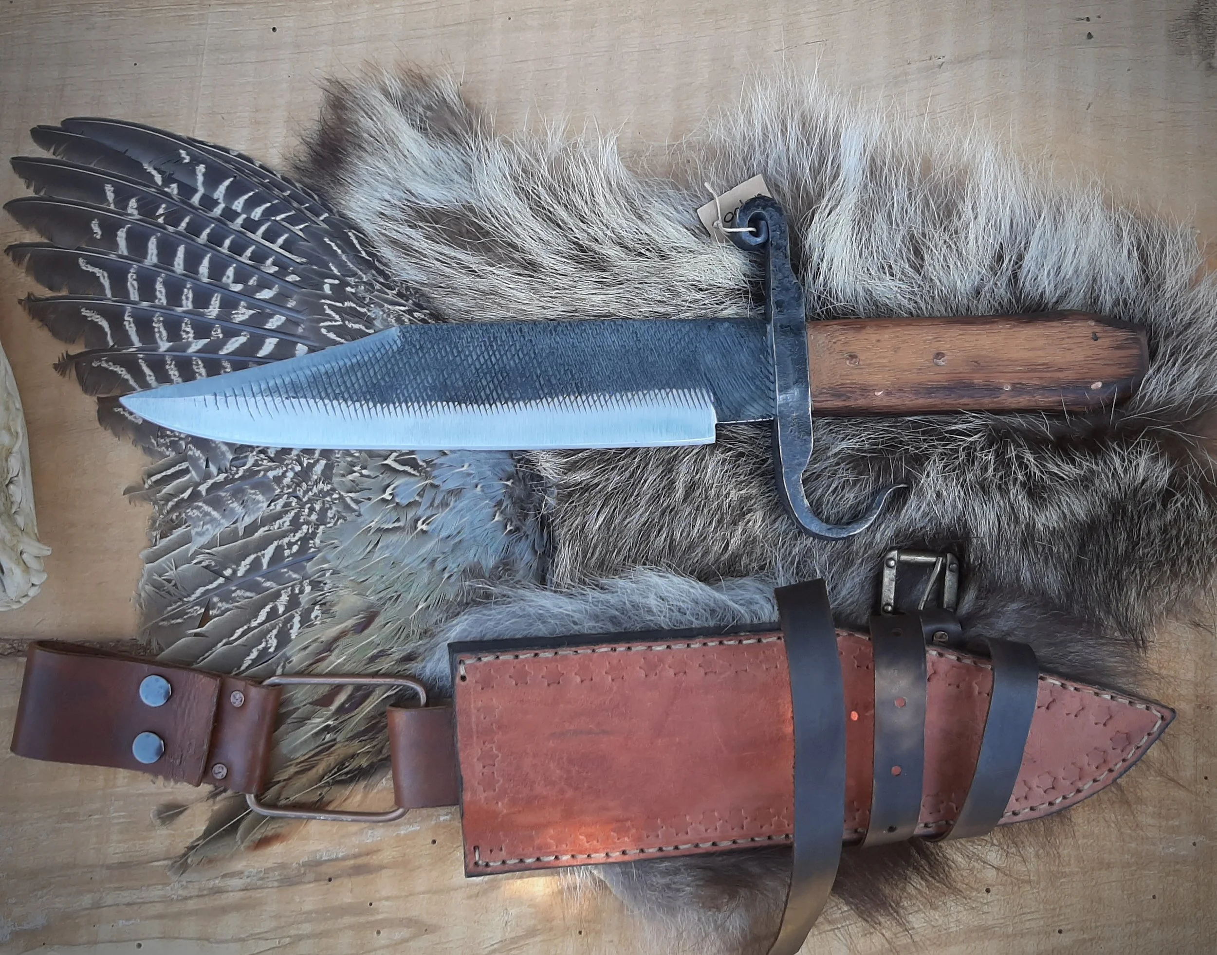 A hunting knife with a leather sheath, feathers, and fur on a wooden surface.