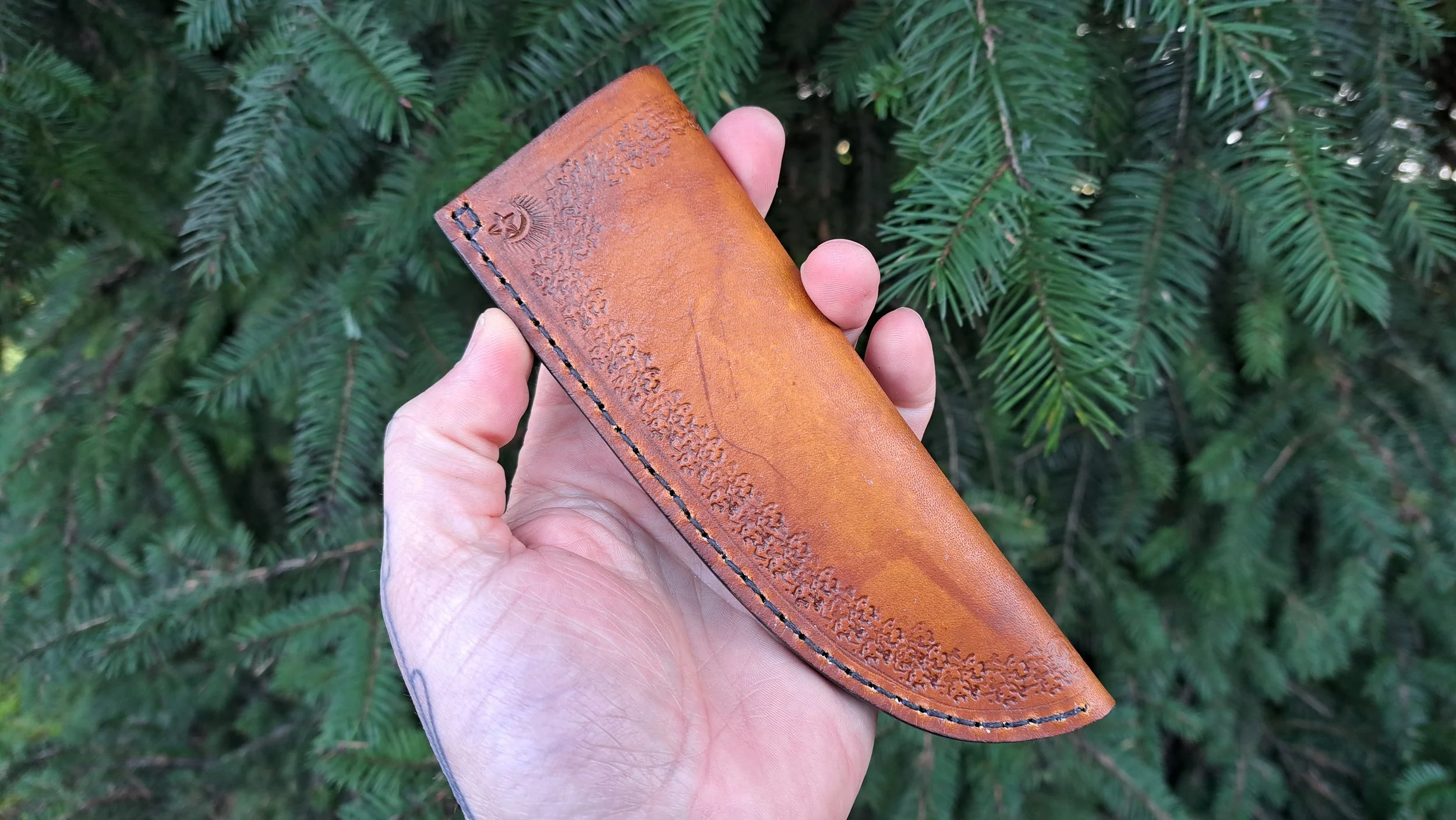 A hand holding a brown leather knife sheath with decorative stitching, in front of green pine tree branches.