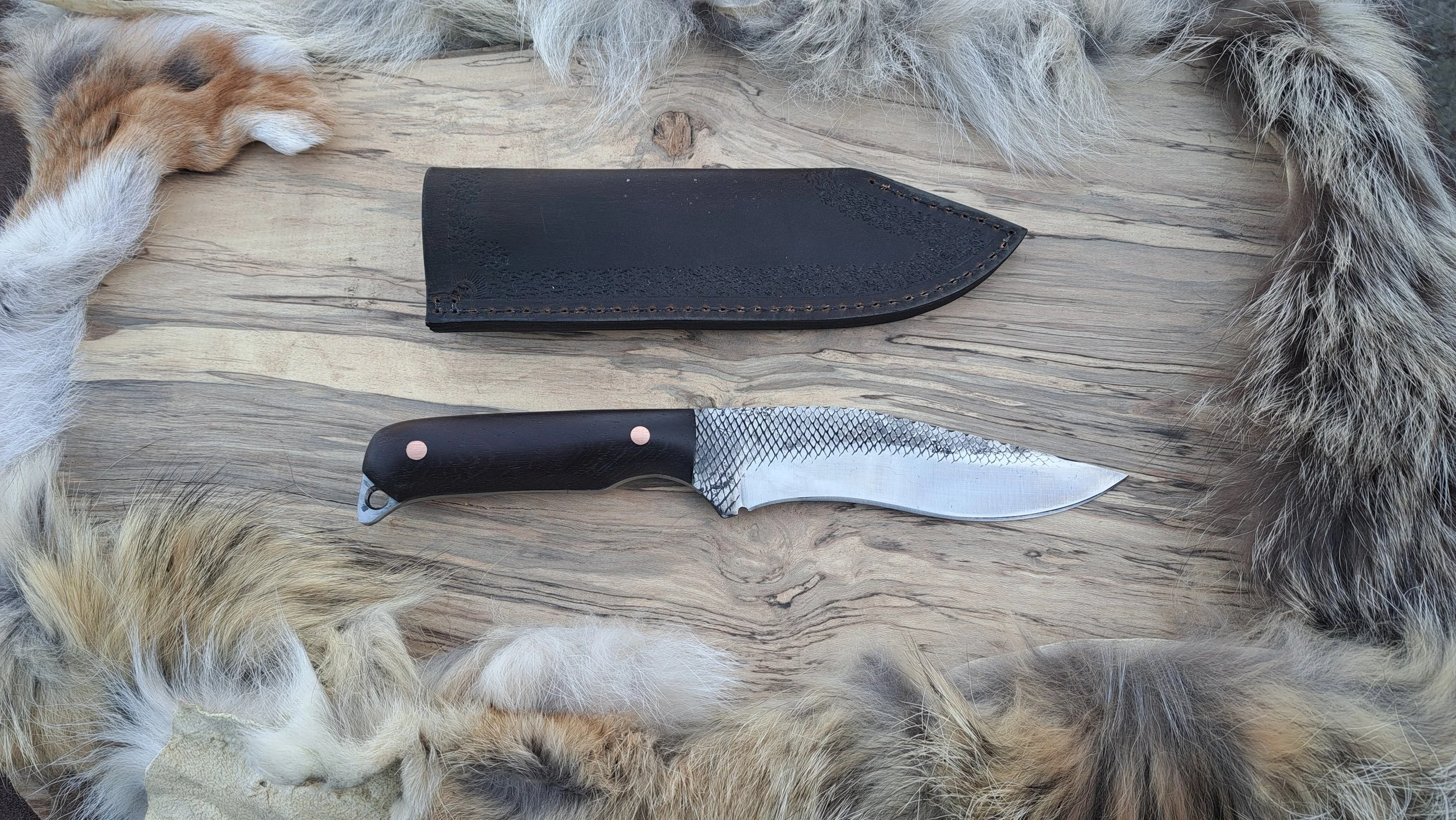 A fixed-blade survival knife with a black handle and a textured silver blade, placed on a wooden surface. The knife has a black leather sheath above it. The scene is framed by the fur of a sleeping animal, possibly a coyote or dog.