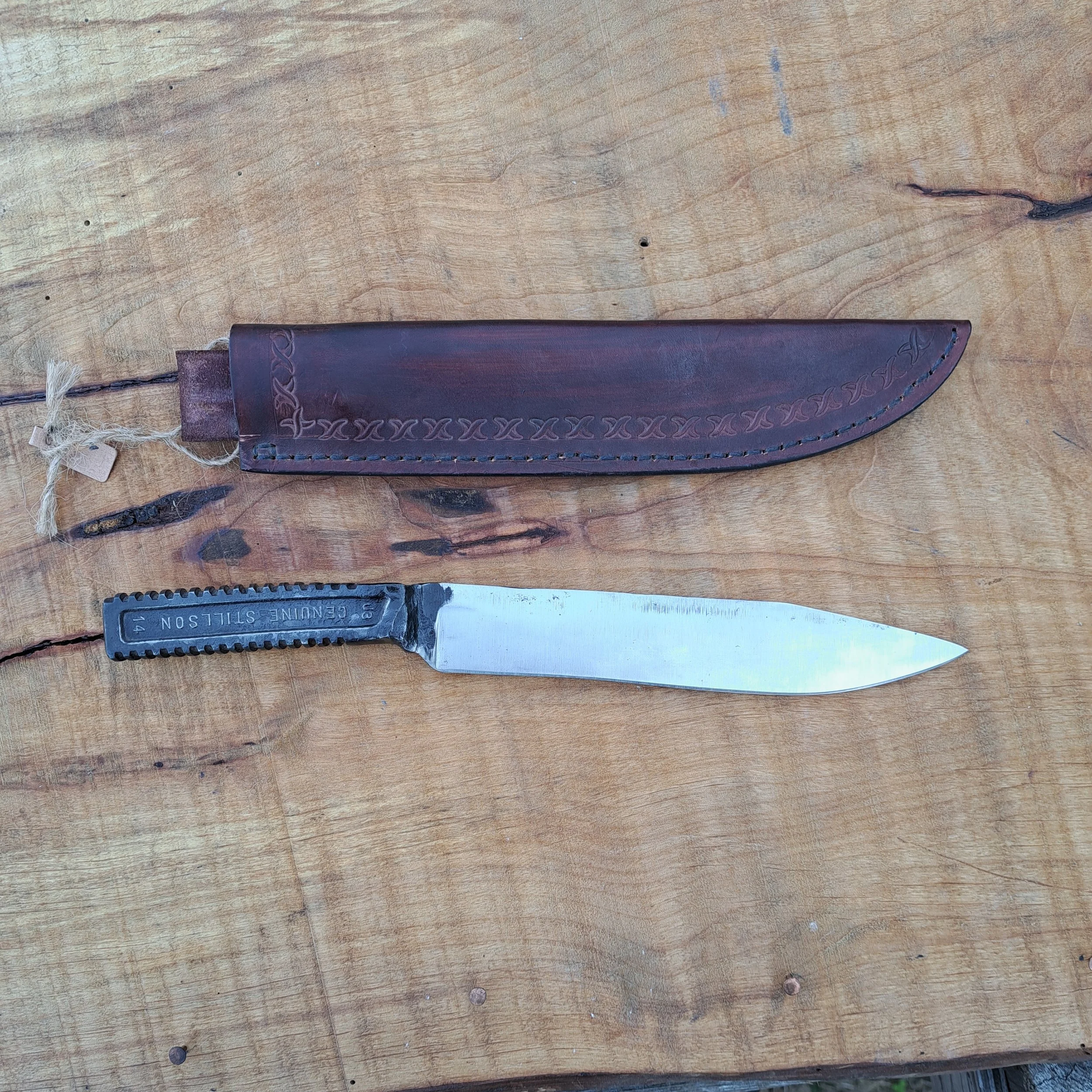 A fixed-blade knife with a black textured handle and a metal blade, placed next to a brown leather sheath on a wooden surface.
