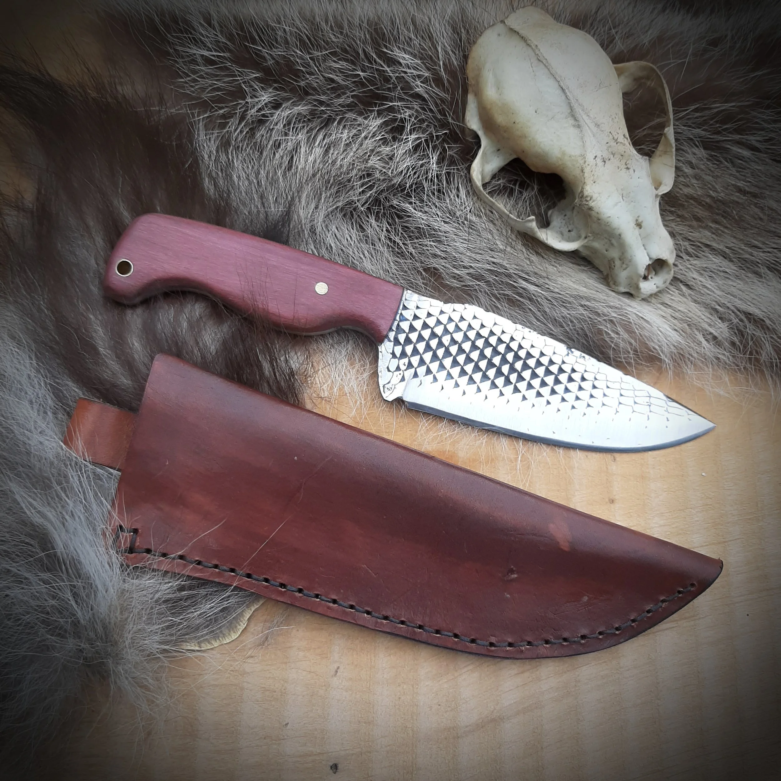 A hunting knife with a textured silver blade, a wooden handle, and a leather sheath, placed on animal fur next to a small animal skull.