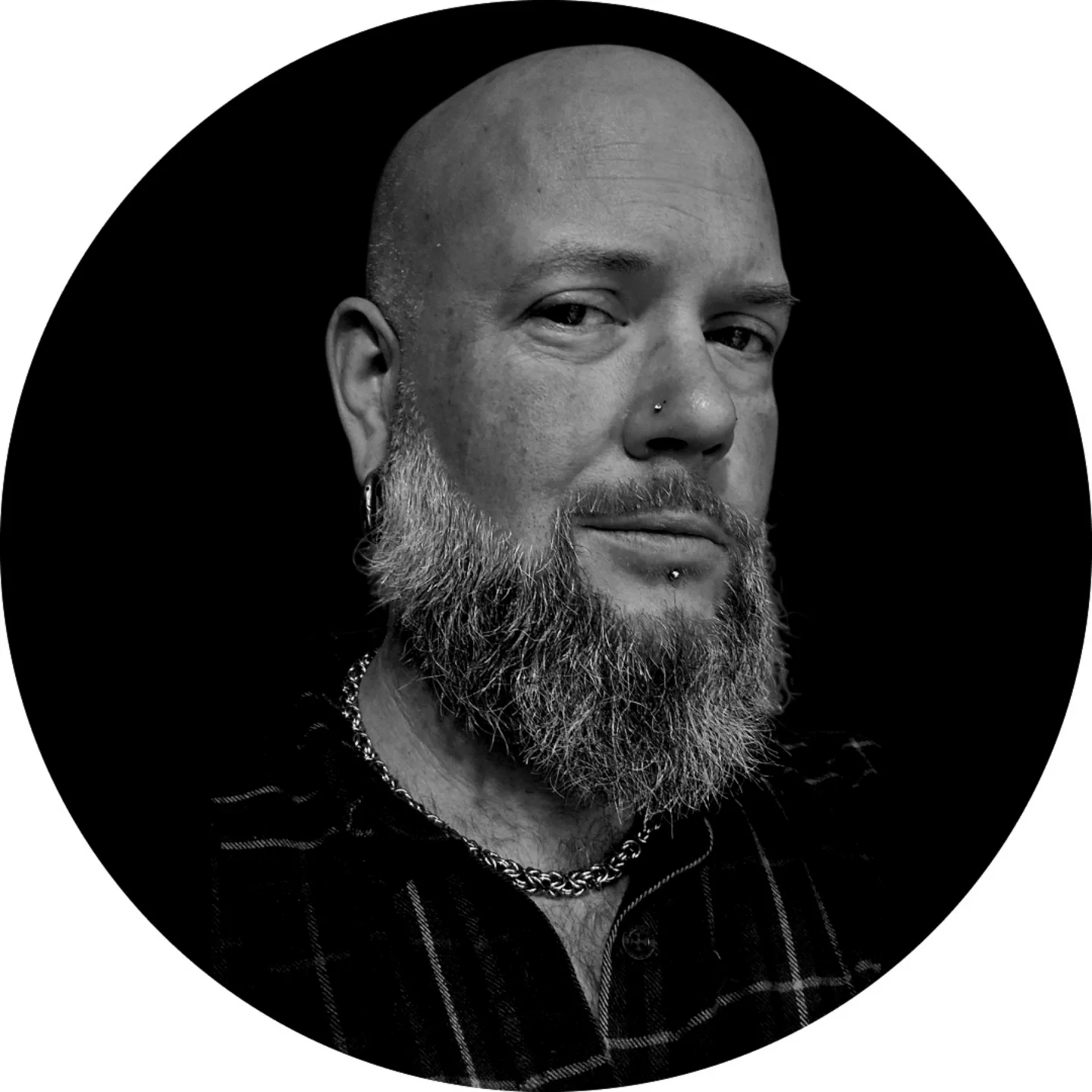 Black and white portrait of a bald man with a full beard, nose piercing, and a chain necklace, wearing a dark plaid shirt, against a dark background.