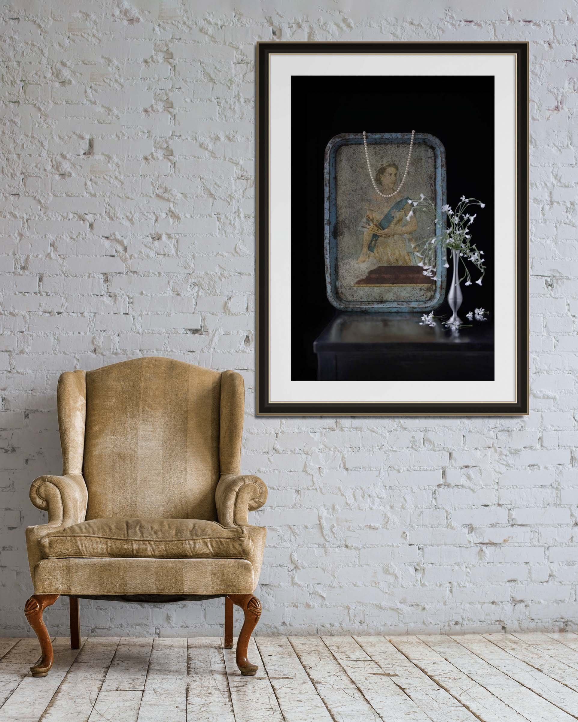 A beige armchair next to a white brick wall with a framed picture hanging above it. The picture shows a vintage portrait with a pearl necklace, a blue tin box, and white flowers in a vase.