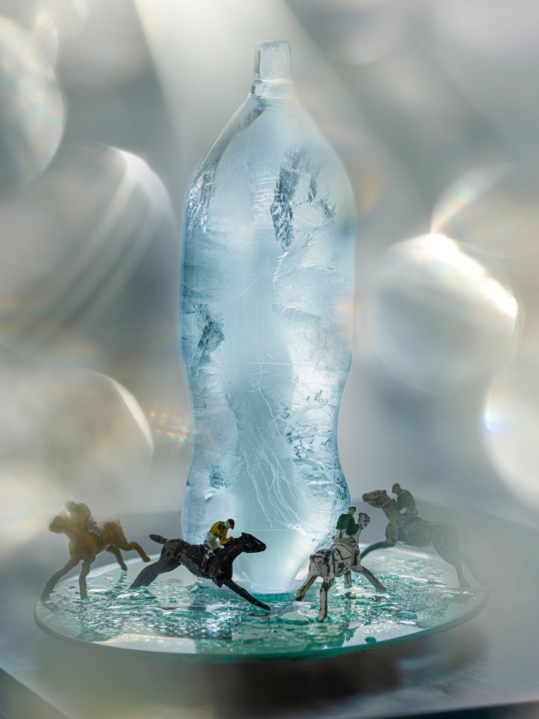 Ice sculpture of a plastic bottle with miniature jockey figurines on a racetrack base.