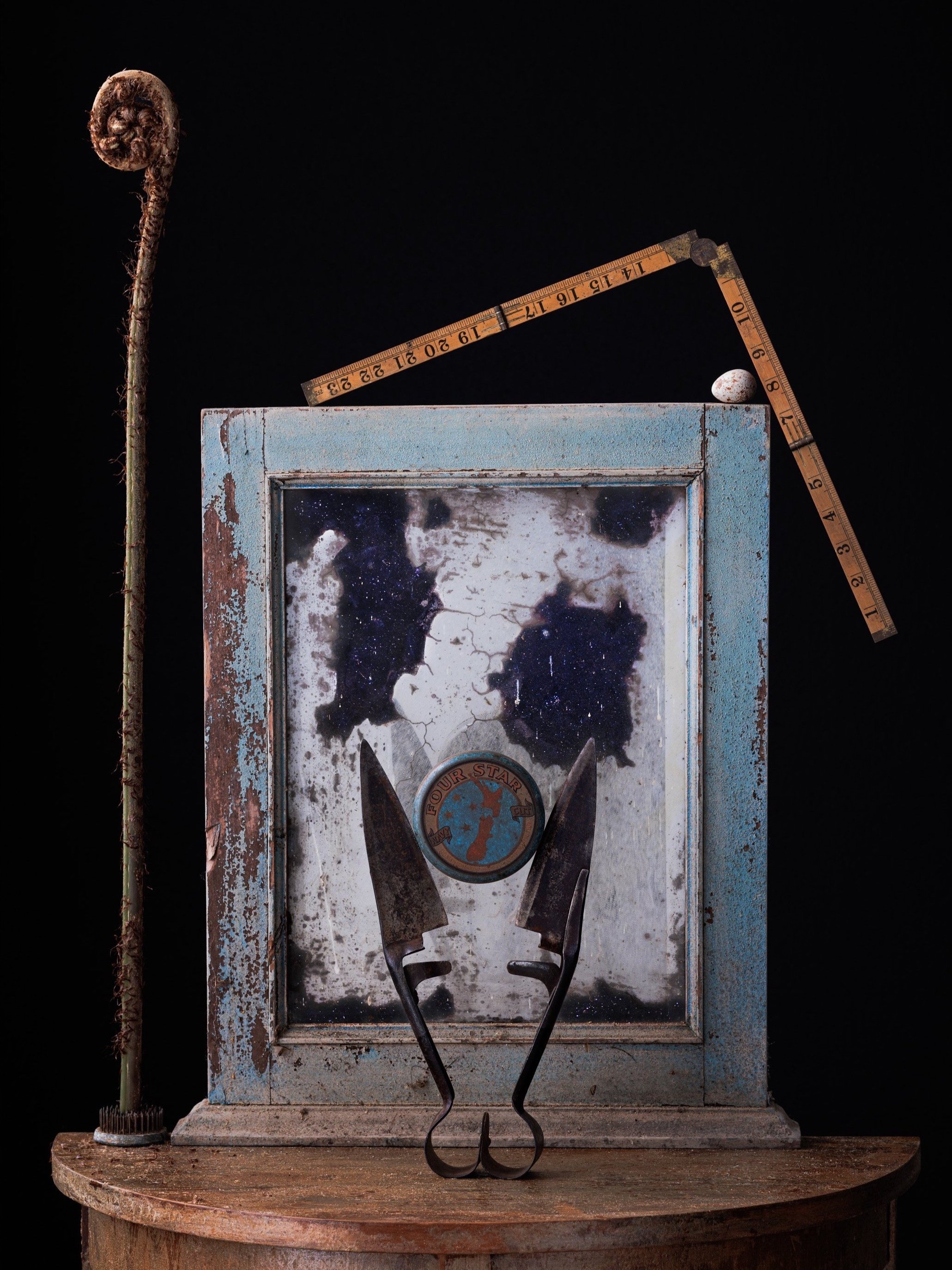 Collection of vintage items including a rusty measuring tape, a weathered blue metal box with a space-themed emblem, two rusted knives, and a tall, dried, curled plant, all arranged on a round wooden surface against a black background.