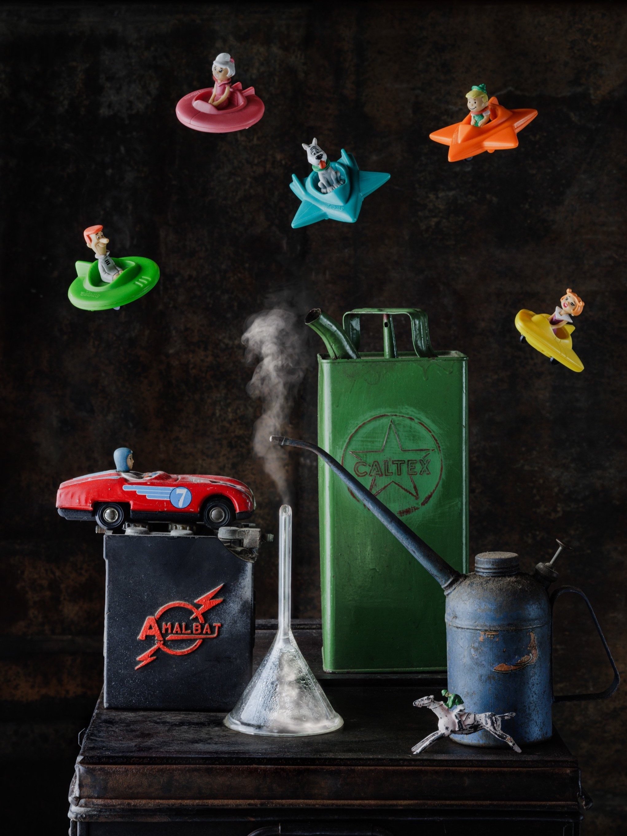 A vintage green Caltex metal gas can, a red toy race car on a black box, a glass flask with a long neck, and a blue metal oil can with a spout on a wooden surface. Several small plastic toys, including characters in colorful floating boats, are suspe