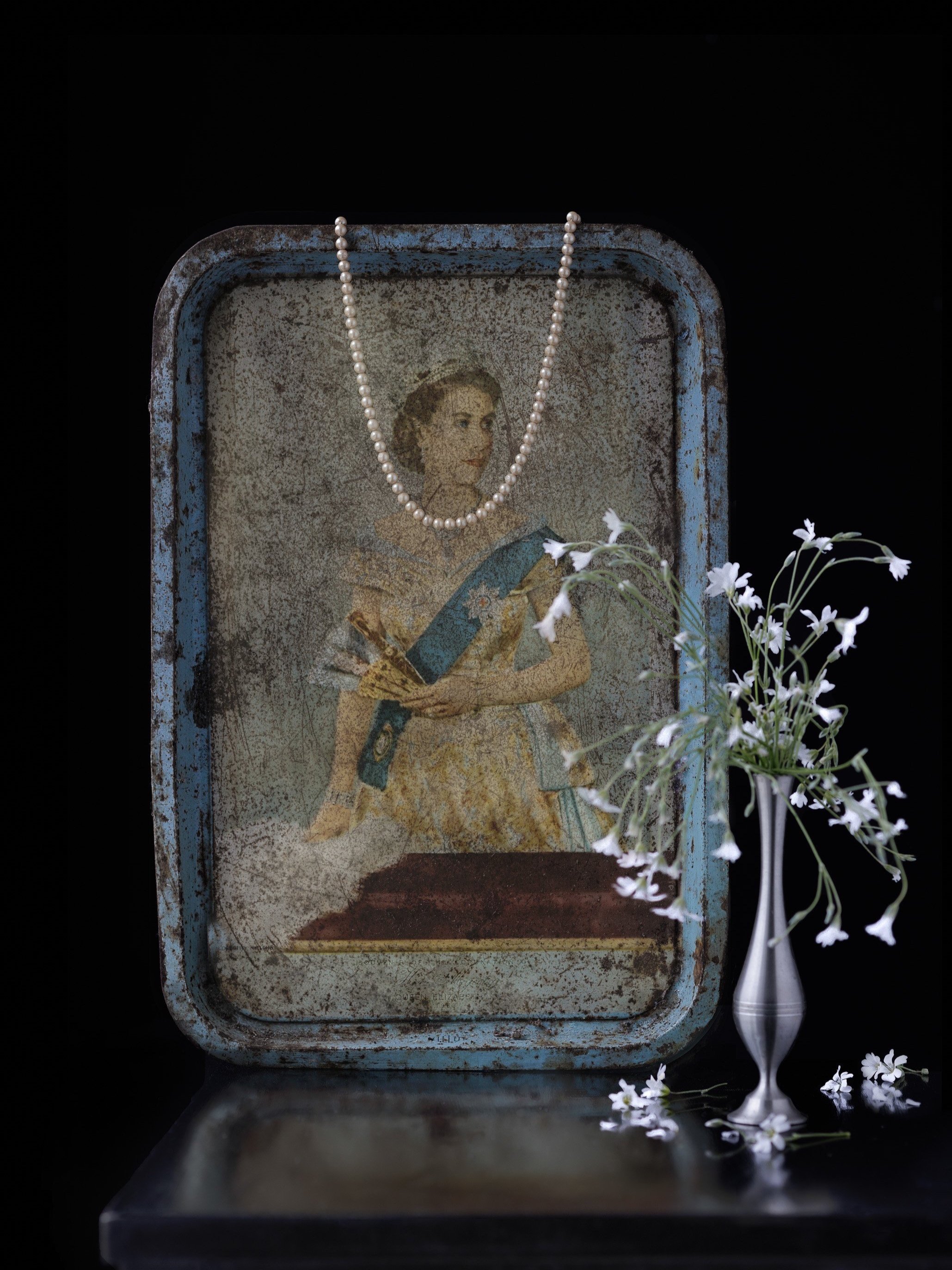 A vintage tin religious icon of a woman, adorned with a pearl necklace, next to a small vase with white flowers, all set against a dark background.