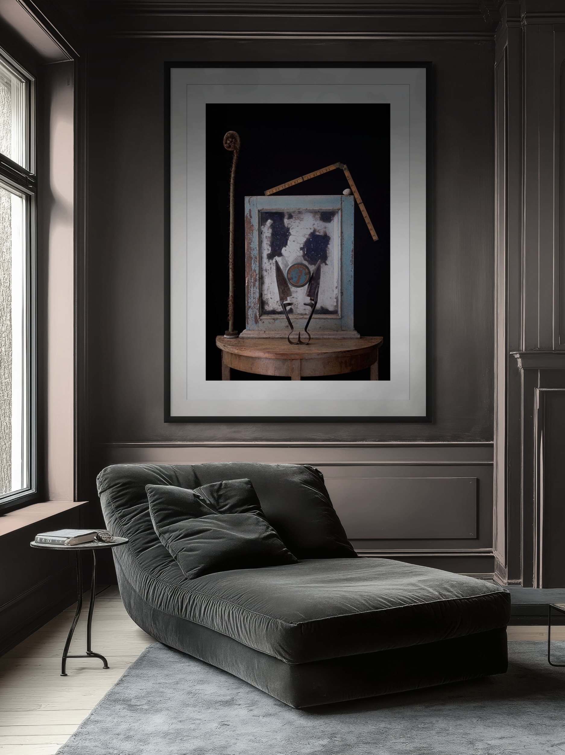 A living room with dark gray walls and paneling, featuring a black chaise lounge with matching pillows, a small side table with a remote and glasses, and a large framed artwork of vintage tools hanging on the wall.