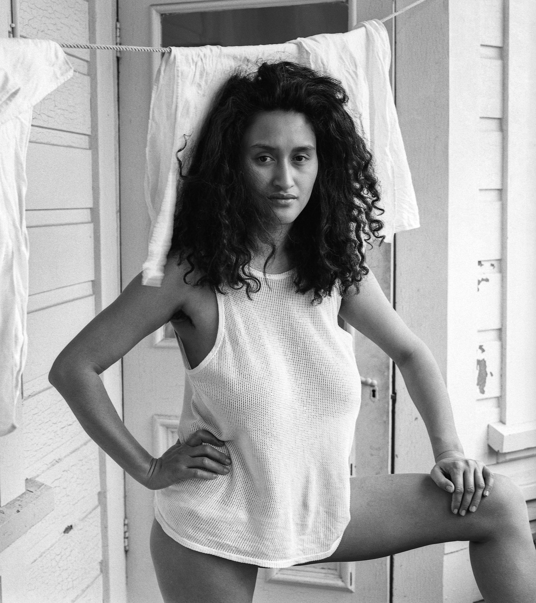 A woman with curly hair in a sleeveless top standing outdoors, with laundry hanging on a clothesline behind her.