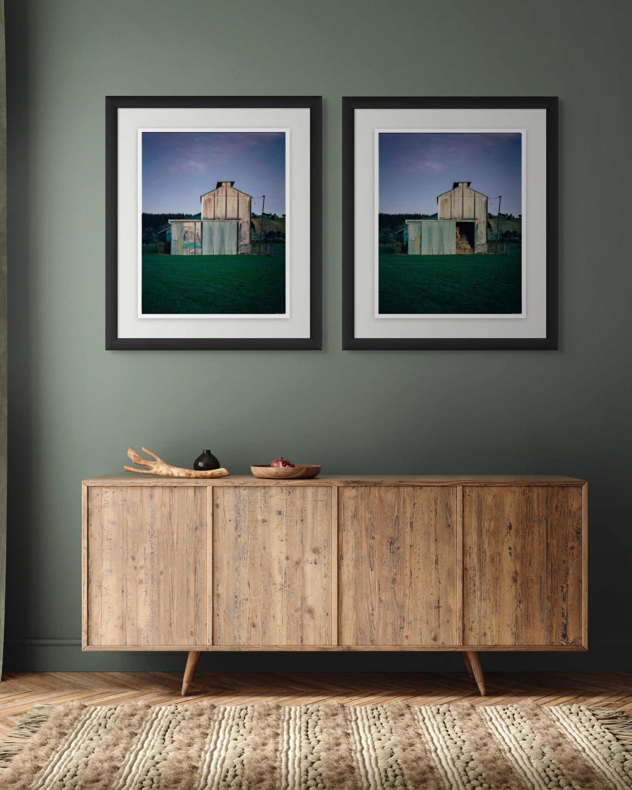 Two framed pictures of a weathered hop kilns hang on a green wall above a light wood sideboard; a decorative branch and a bowl with red fruit sit on the sideboard, with a striped beige rug on the wooden floor.