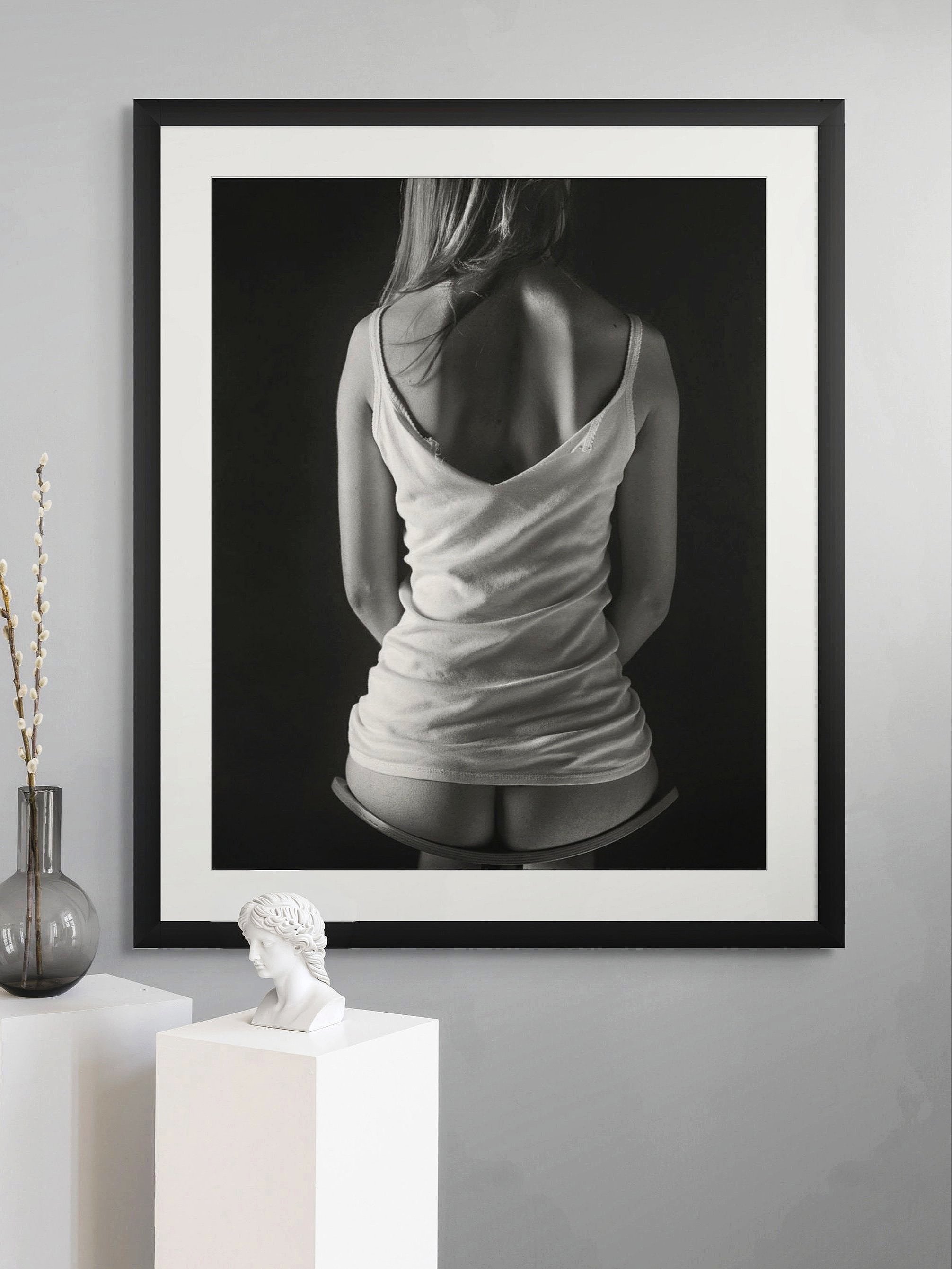 Black and white photograph of a woman with her back to the camera, wearing  just a torn singlet, displayed in a black frame on a gray wall.