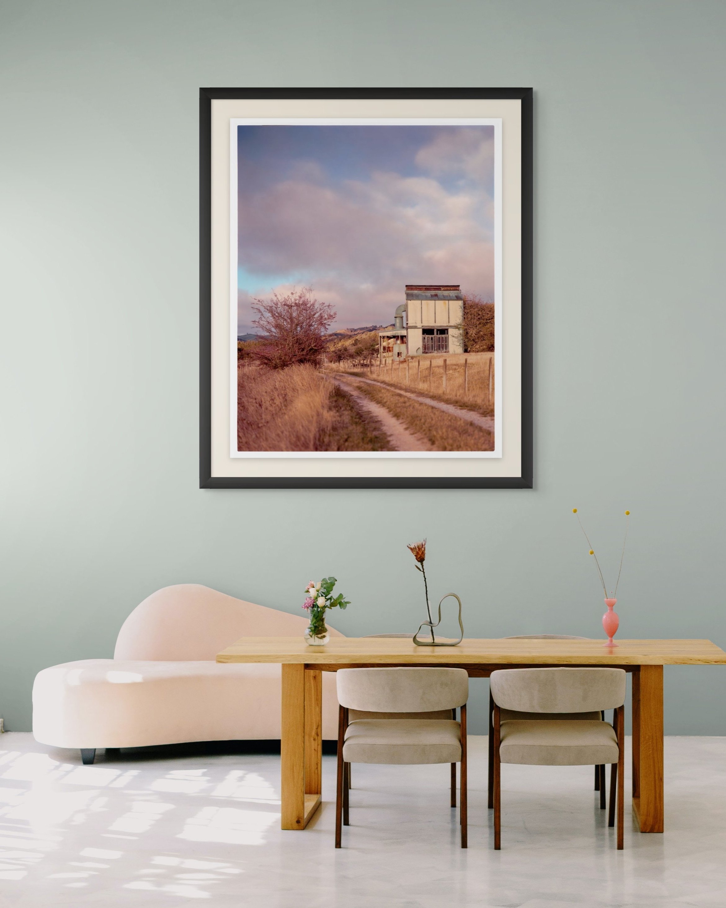 A framed landscape photograph of a dirt path leading to an old hop kiln in a rural area hangs on a light green wall in a modern living room. 