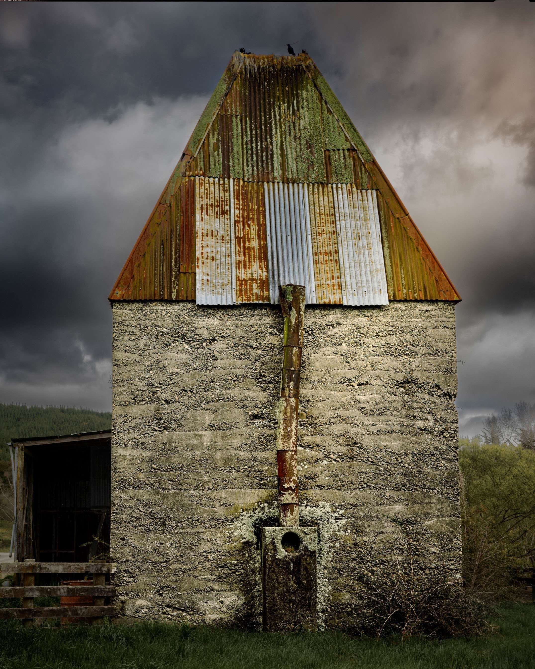 An old, weathered concrete hop kiln with a stone foundation and a rusted corrugated metal roof under a dark, stormy sky.