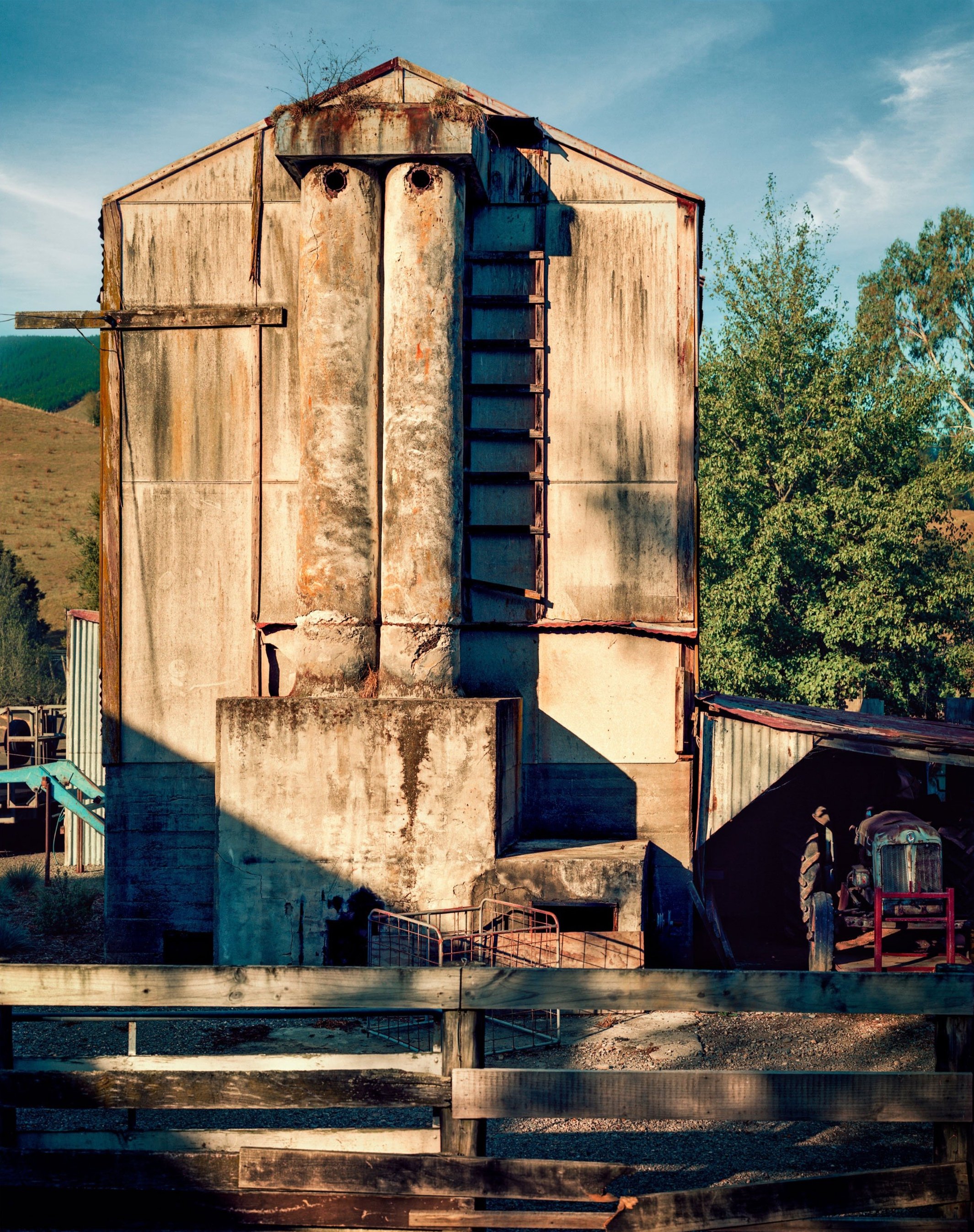 Old, concrete hop kiln, with a ladder on the front and a pasture fence in the foreground. A tractor is parked inside a shed on the lower right, with a cloudy sky and trees in the background.