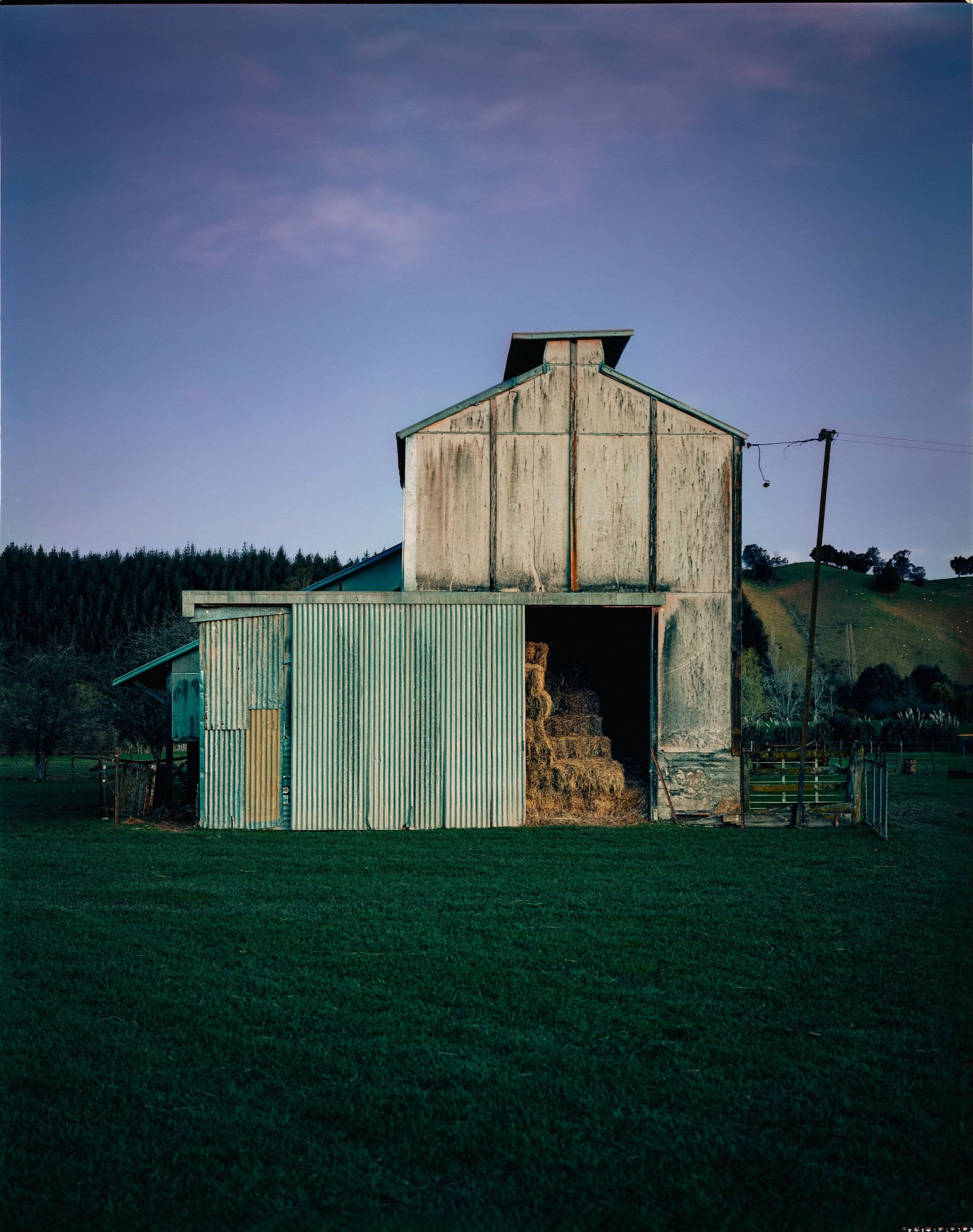 A rustic farm hop kilns with hay bales inside, set against a rural landscape at dusk, with a green grassy field in the foreground and rolling hills in the background.