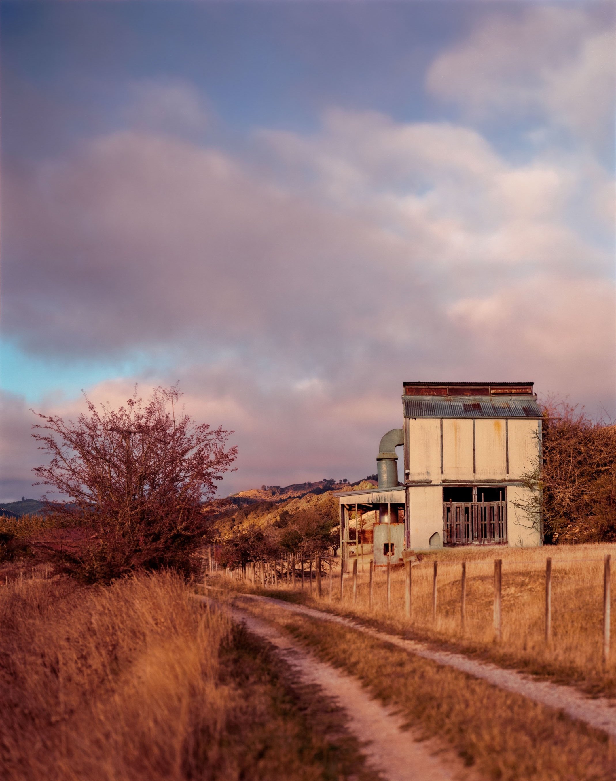An old, weathered hop kiln with a rusted metal roof and a large chimney, surrounded by a dirt path, a wire fence, and trees in a rural landscape under a partly cloudy sky during sunset.