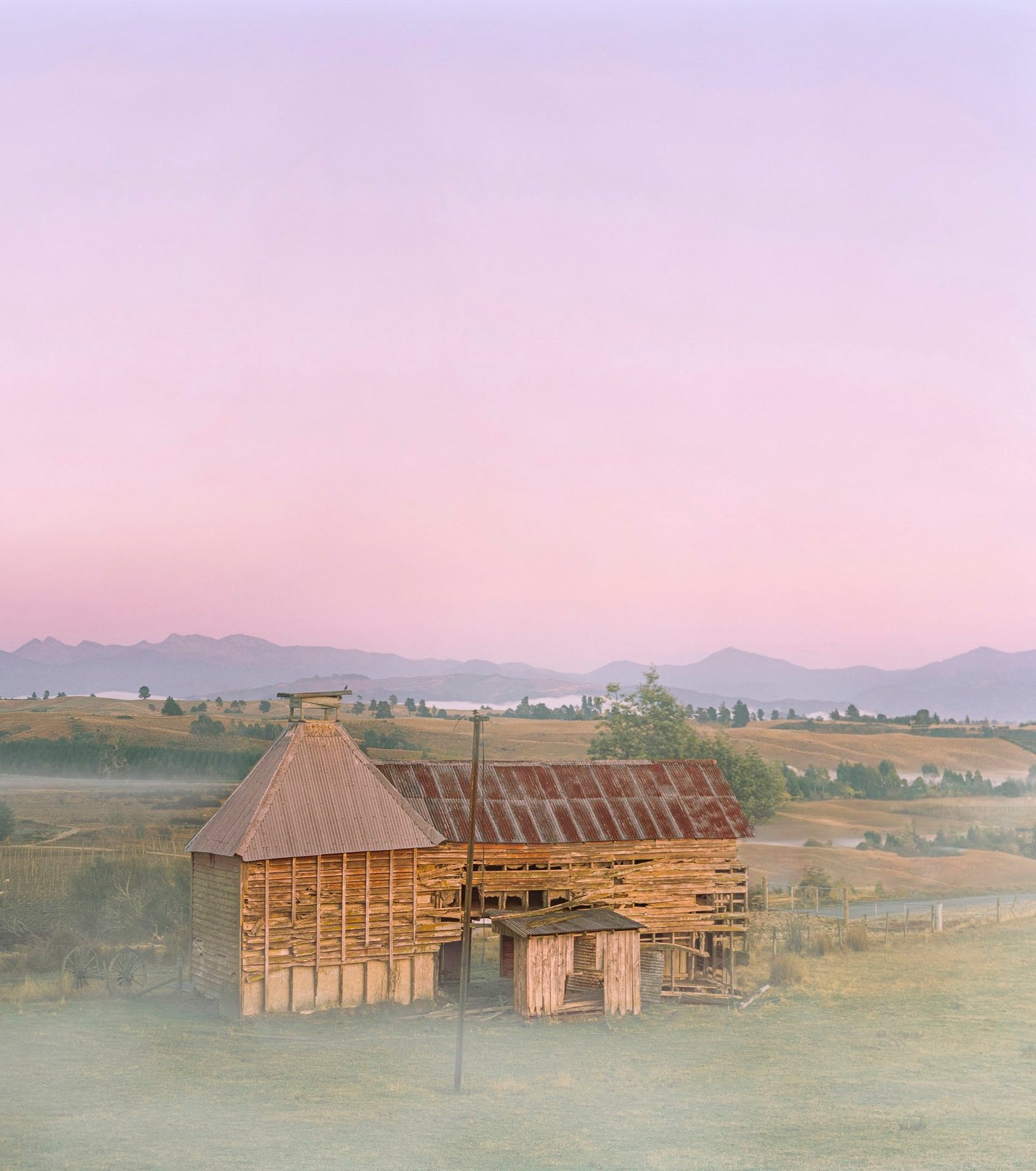 A rustic wooden hop kiln with a corrugated metal roof in a rural landscape during dawn, with mountains in the distance and a pink and purple sky.