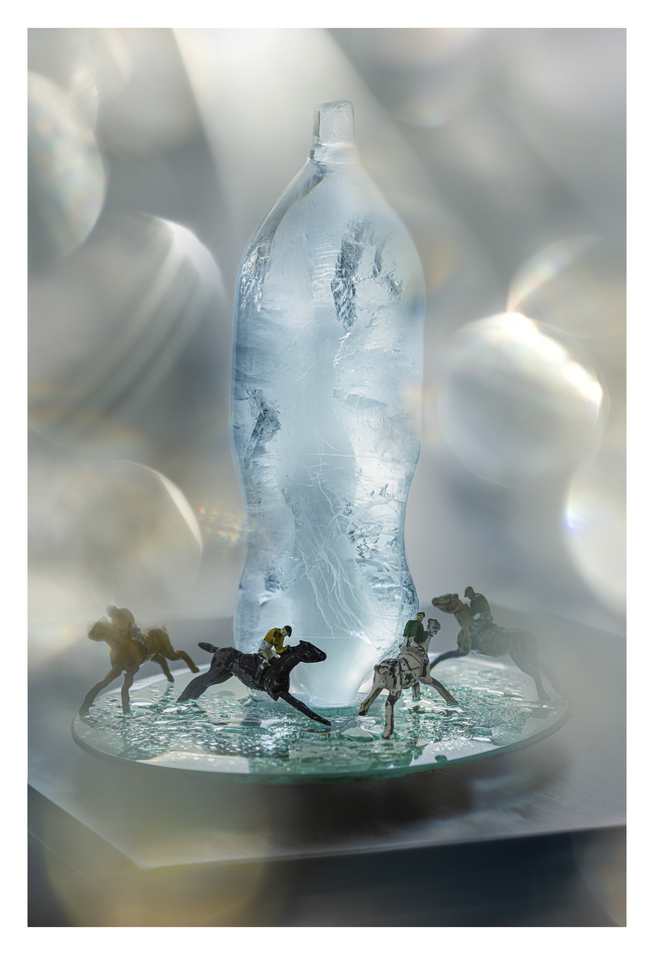 A glass sculpture of a water bottle with miniature horse figurines and jockeys racing around a circular base in front of the bottle, with a blurred, sparkling background.