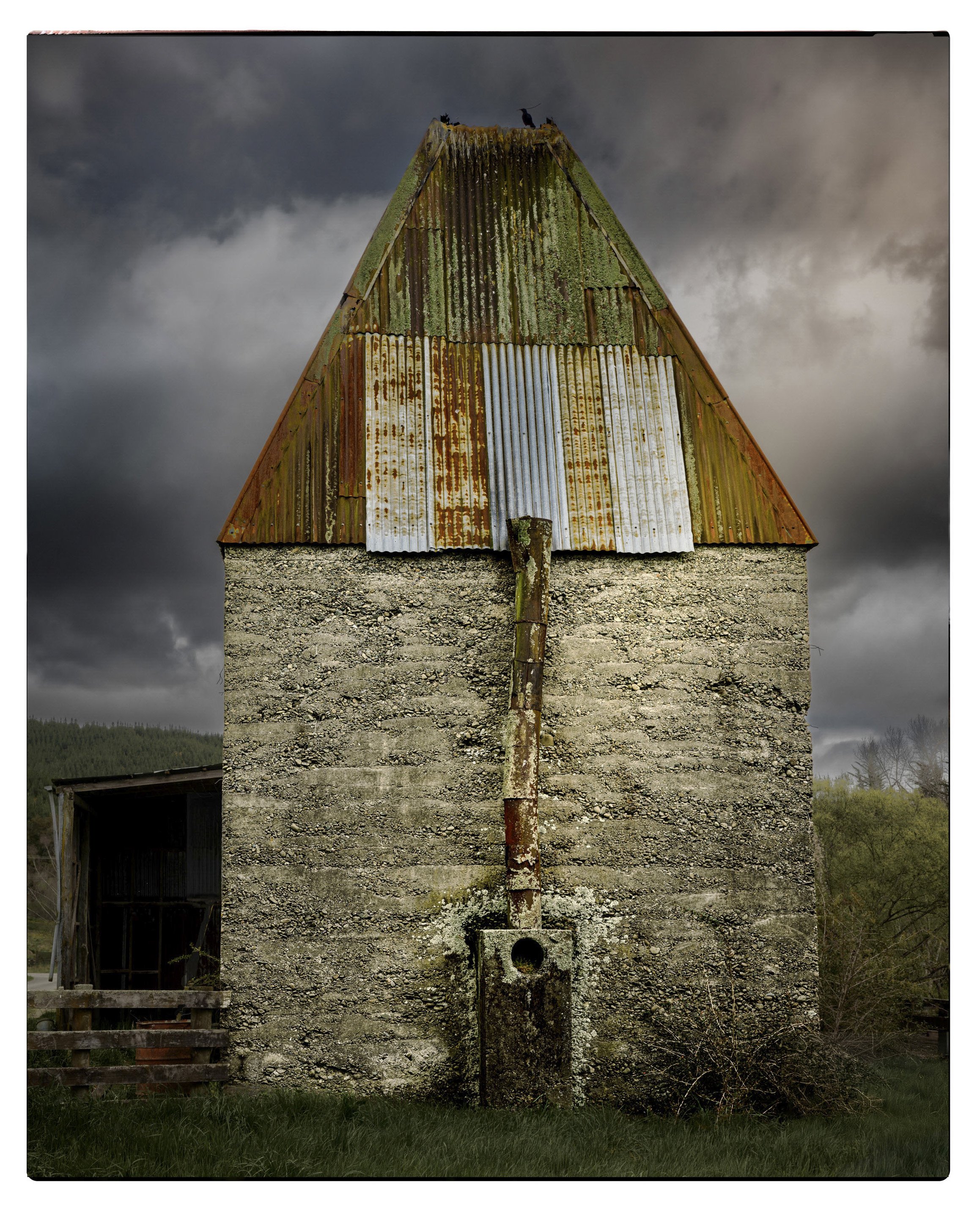 An old, weathered stone building with a rusty, patchwork metal roof. There is a rusty pipe running down the front of the building and a small concrete structure at the base. The sky is overcast with dark clouds.
