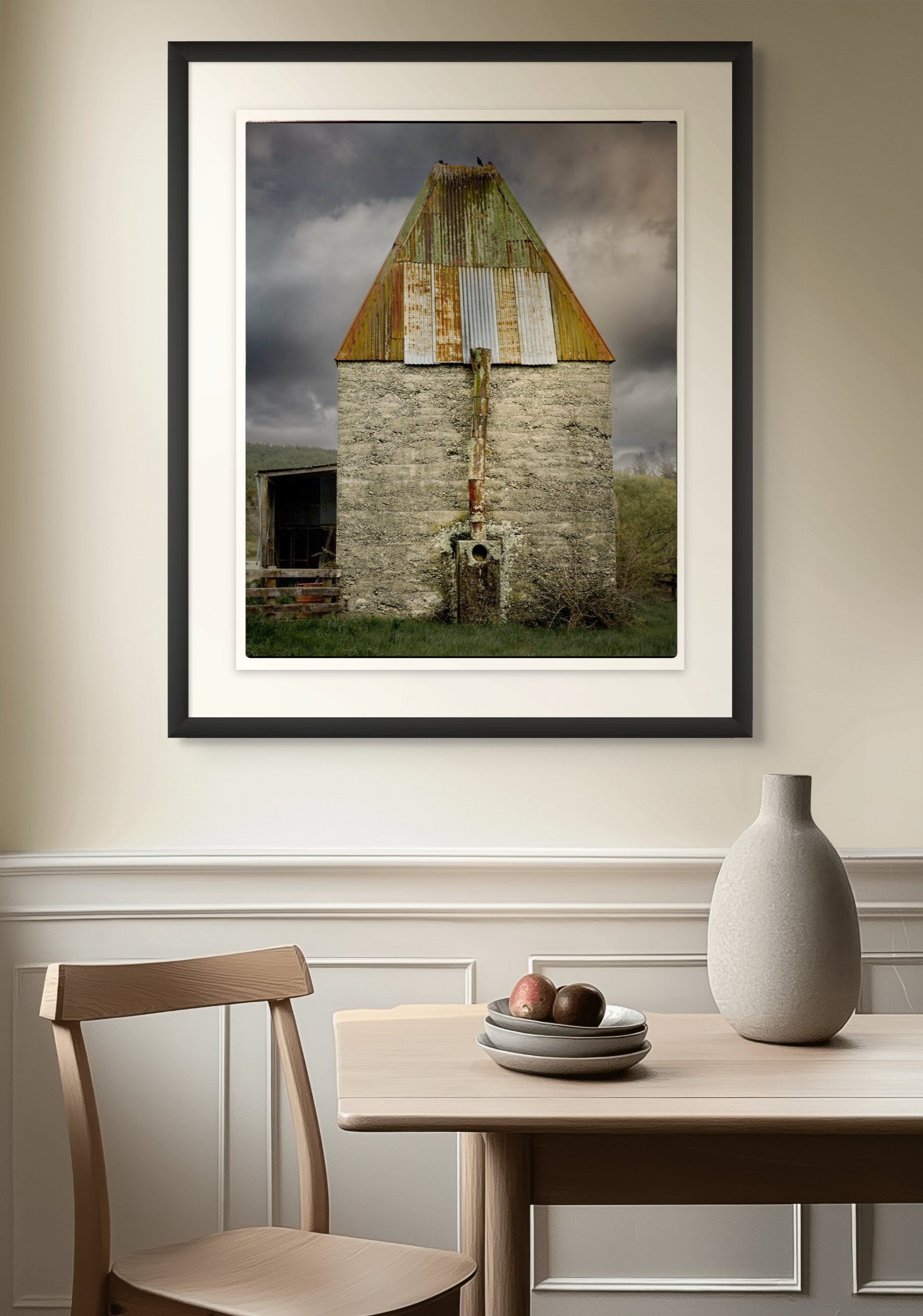 Framed photograph of an old, weathered hop kiln with a rusty metal roof, hanging on a beige wall above a dining table with a white bowl of fruit and a beige vase.