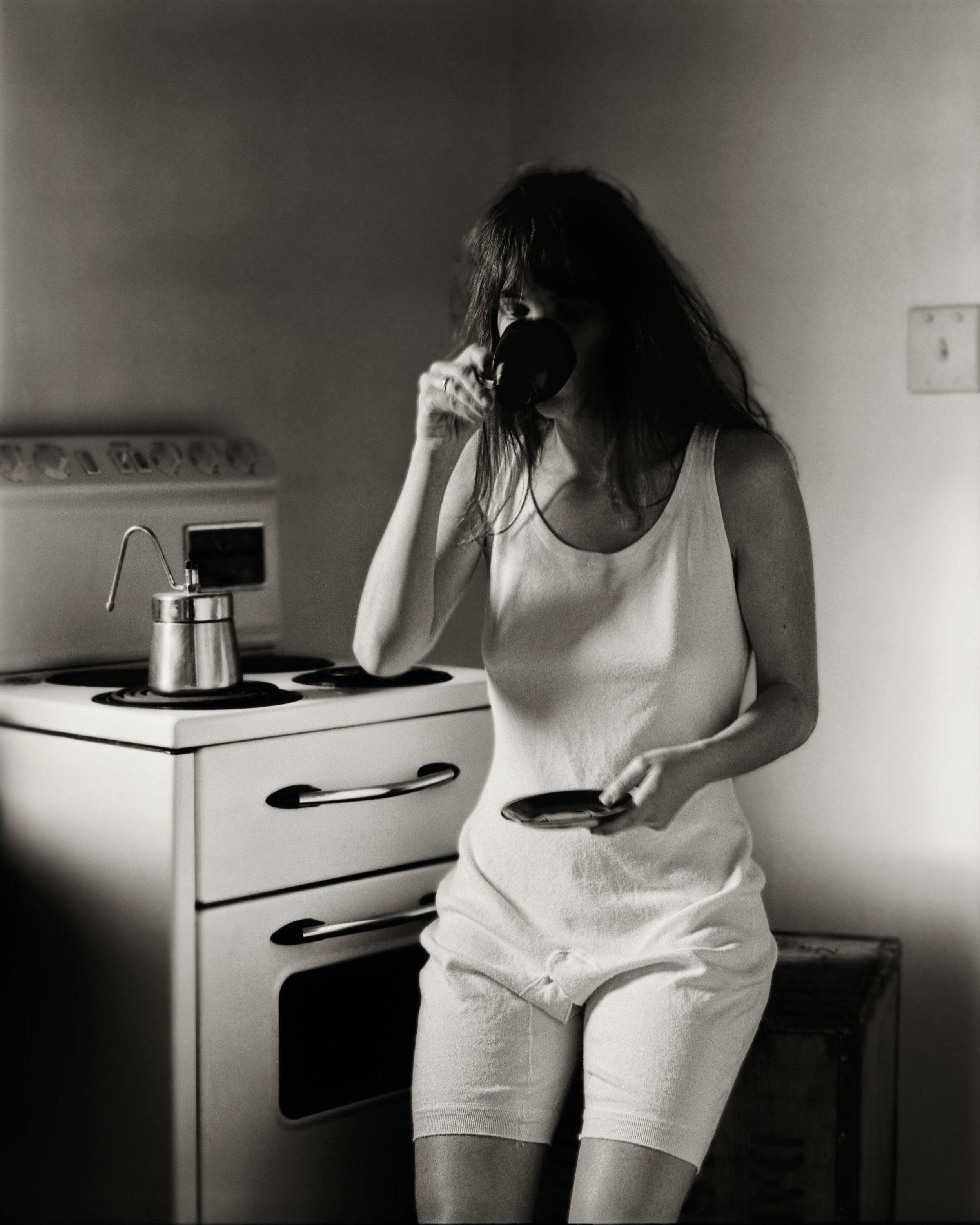 A woman wearing a mans all in one undergarment, drinking from a cup while standing beside a stove in a dimly lit kitchen.