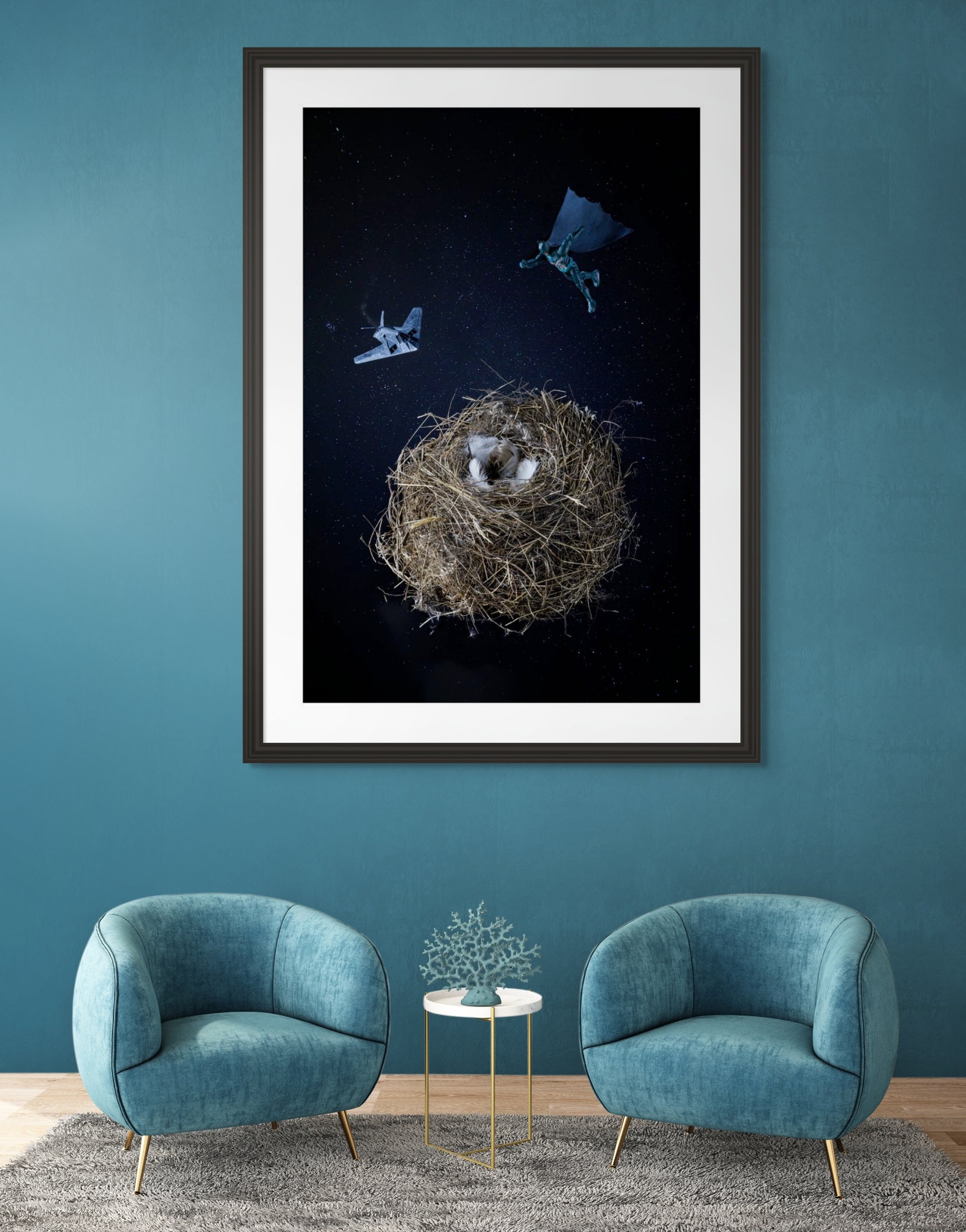 A framed spaceship scene with a bird's nest at the center, a small bird inside the nest, a space shuttle, and a flying figure in a cape against a starry sky, hanging on a teal wall in a living room with two blue chairs and a small side table.