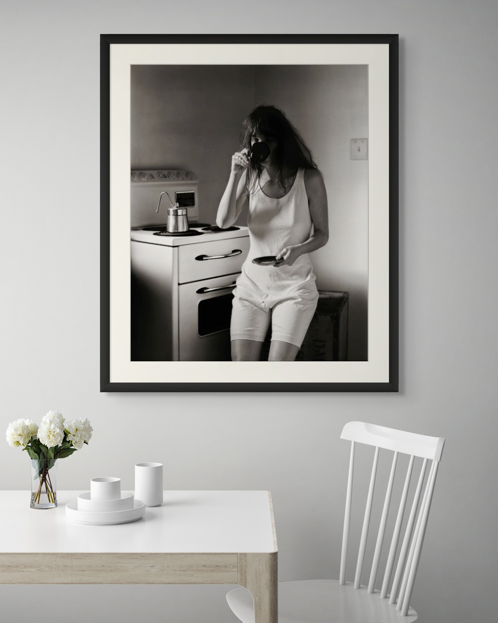 A black and white photograph of a woman in a kitchen, drinking from a cup, holding a plate, standing beside a stove.