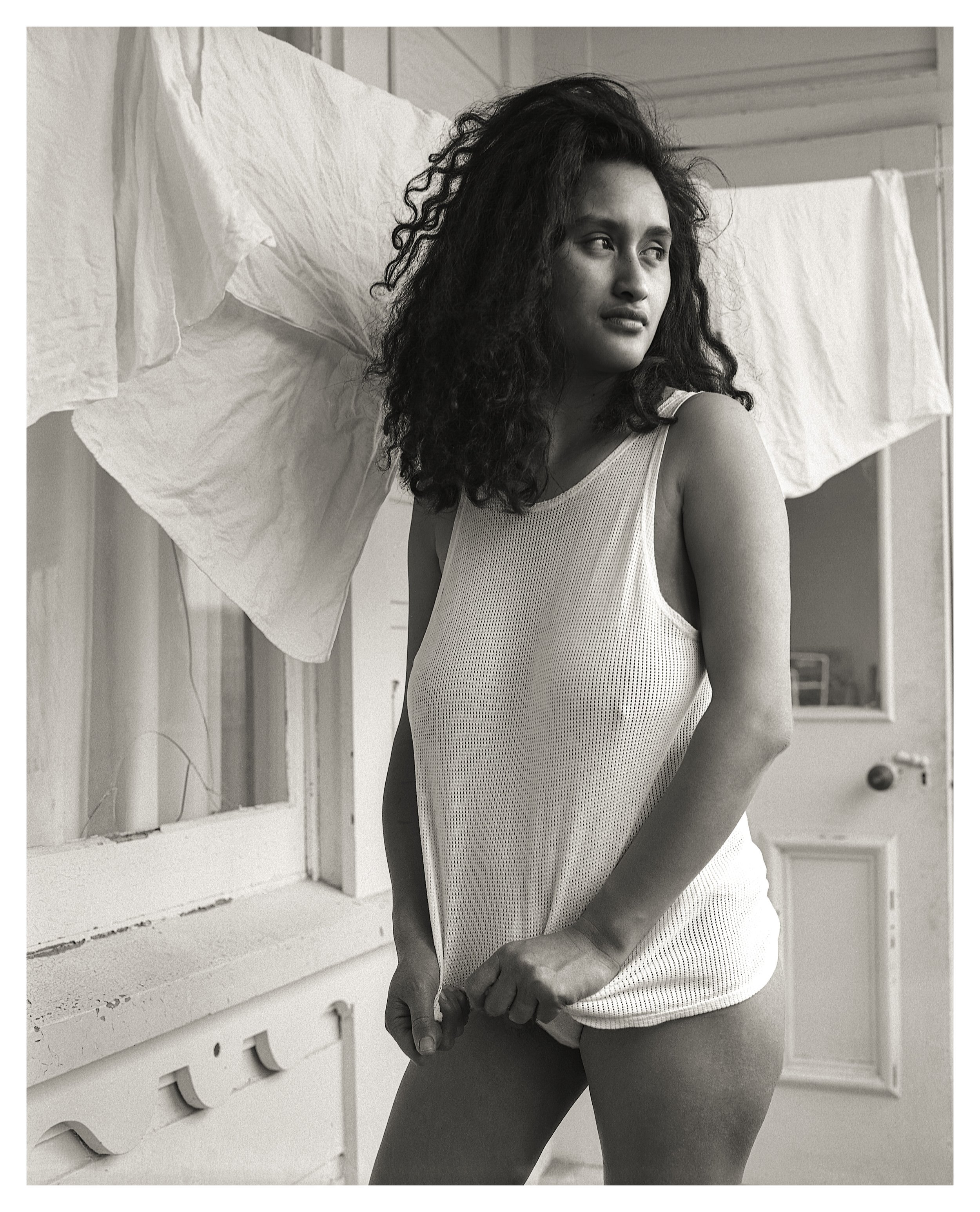 A woman with curly hair in a sleeveless top standing indoors with laundry hanging in the background.