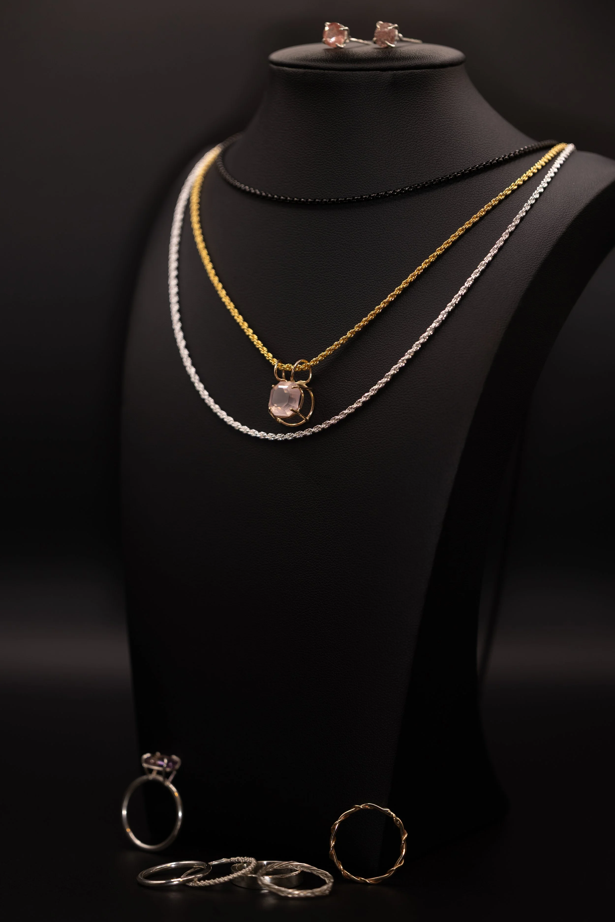 Display of jewelry on a black bust, including layered necklaces with one featuring a large gemstone pendant, and rings on a black surface, against a dark background.