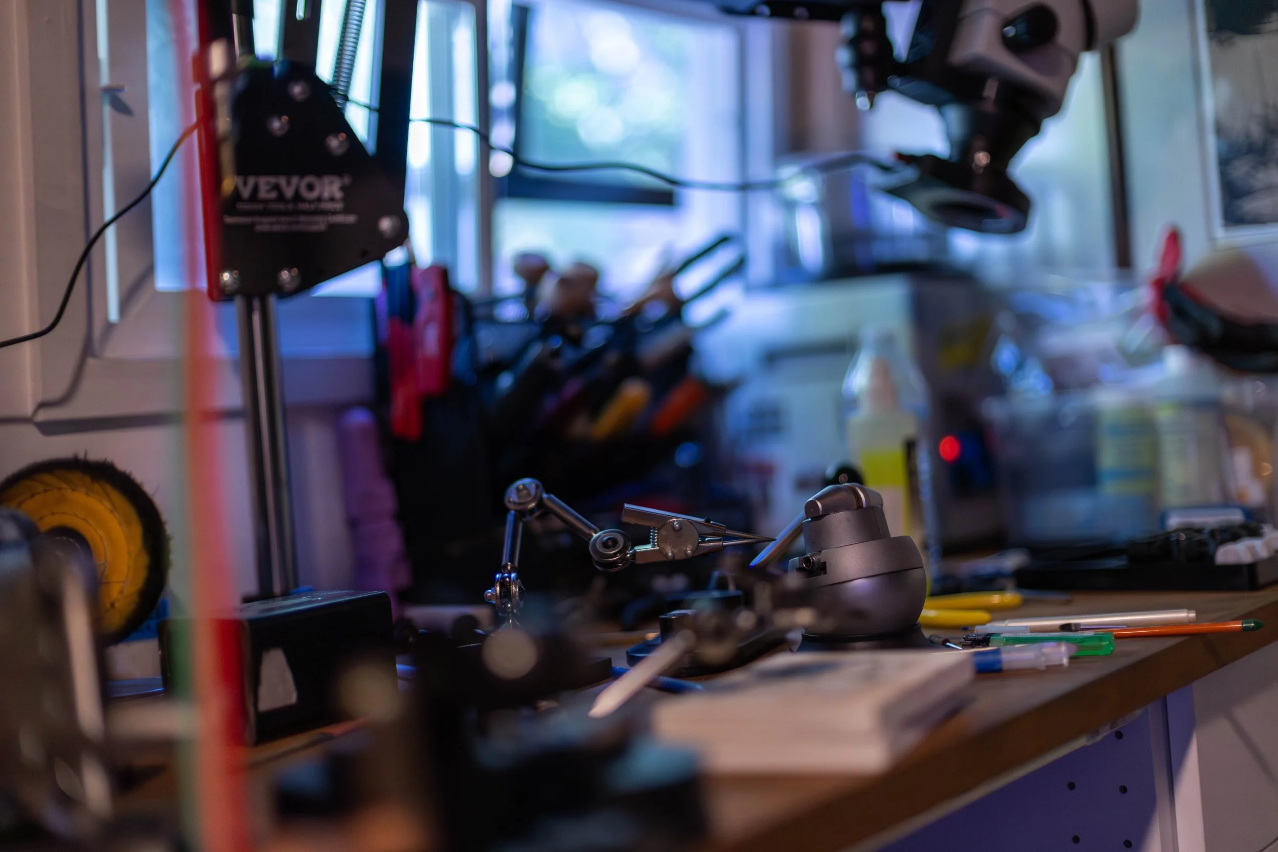 A cluttered jewelry workbench filled with tools and equipment, including a microscope, vise, and various metalsmithing hand tools, with a window in the background.