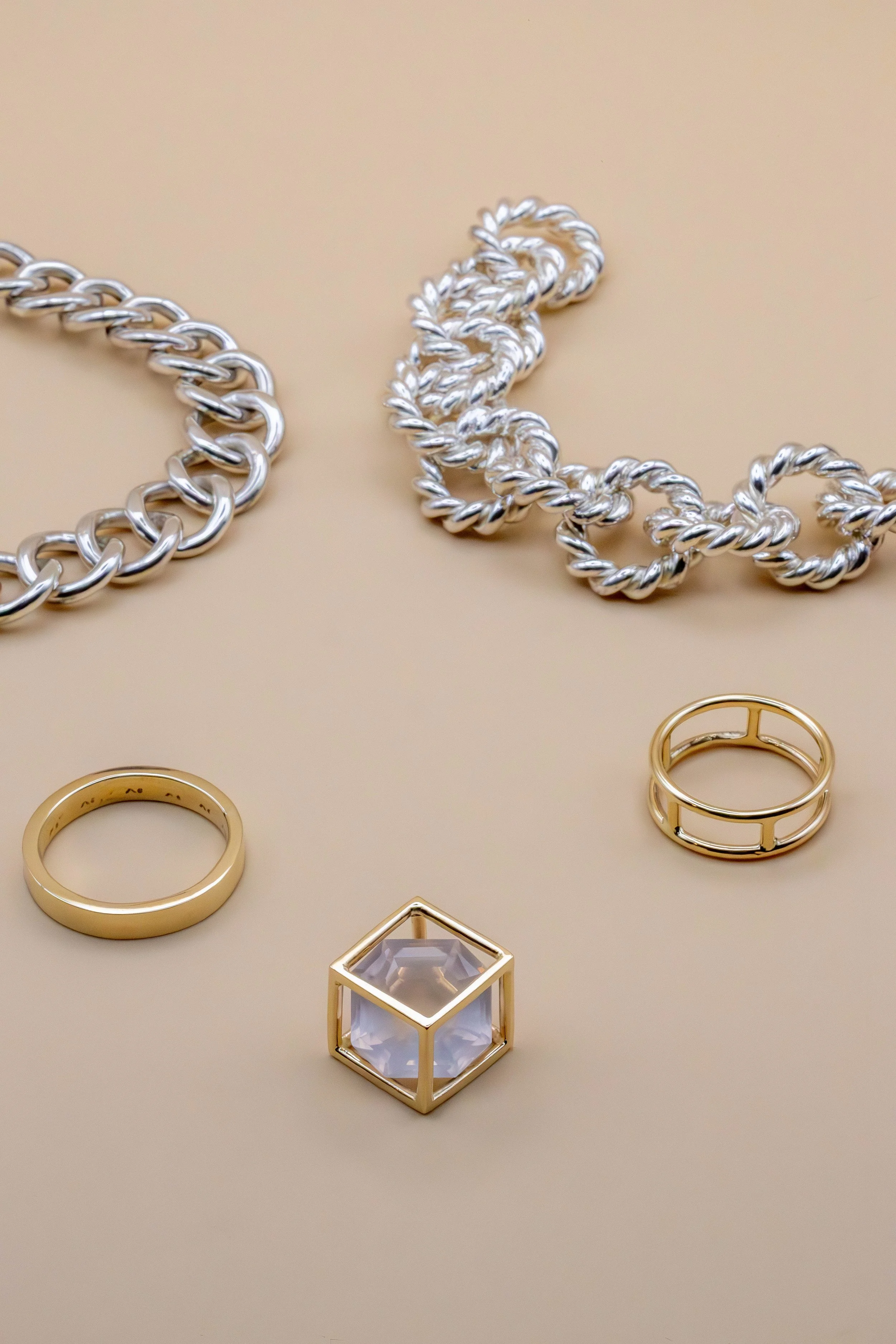 Silver and gold jewelry pieces, including silver chain necklaces, gold rings, and a golden cube on a beige background.