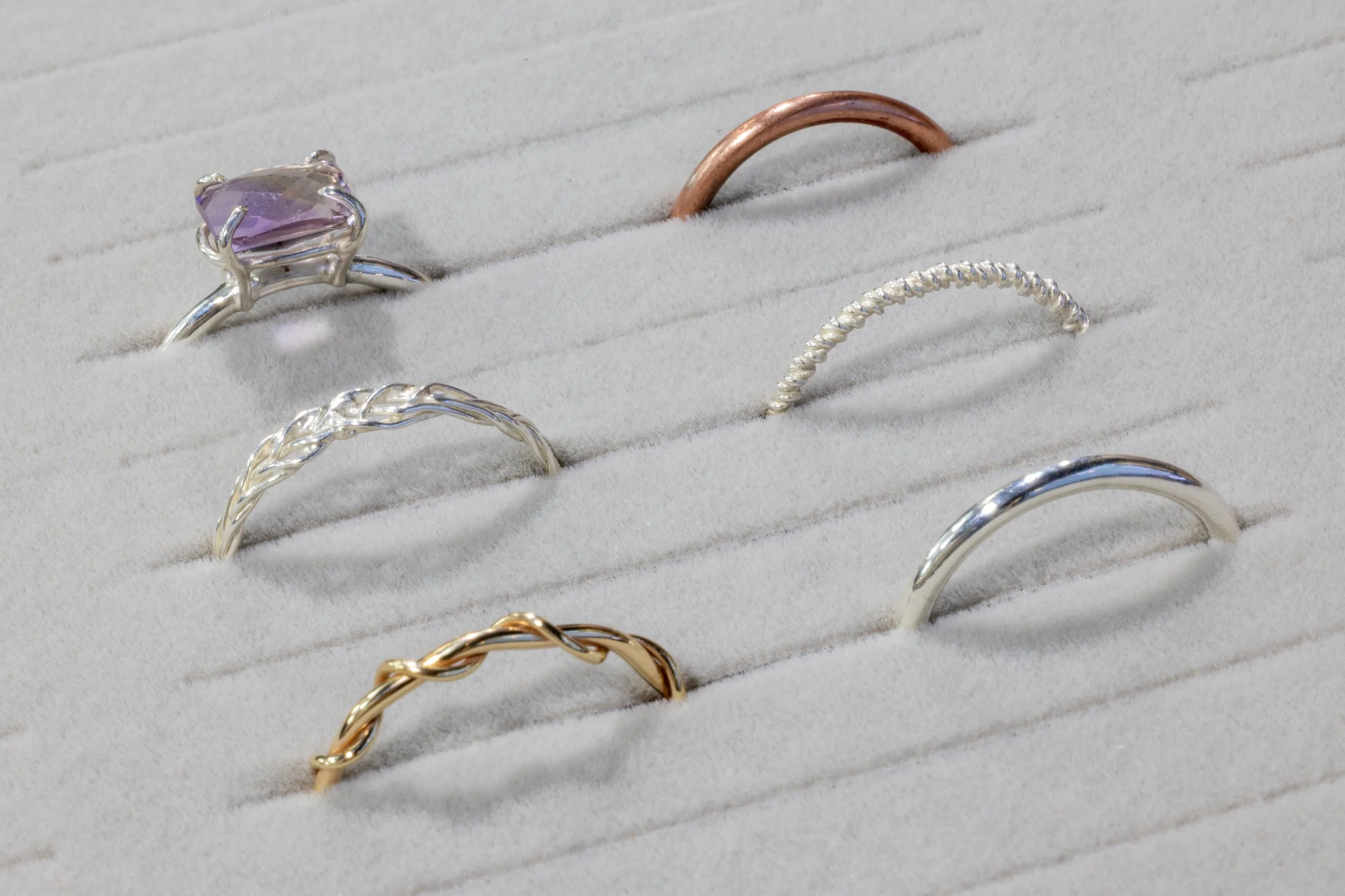 Display of various rings on a white padded surface, including a silver ring with a purple gemstone, a rose gold band, a silver band with a twisted design, a silver textured band, and a gold twisted band.