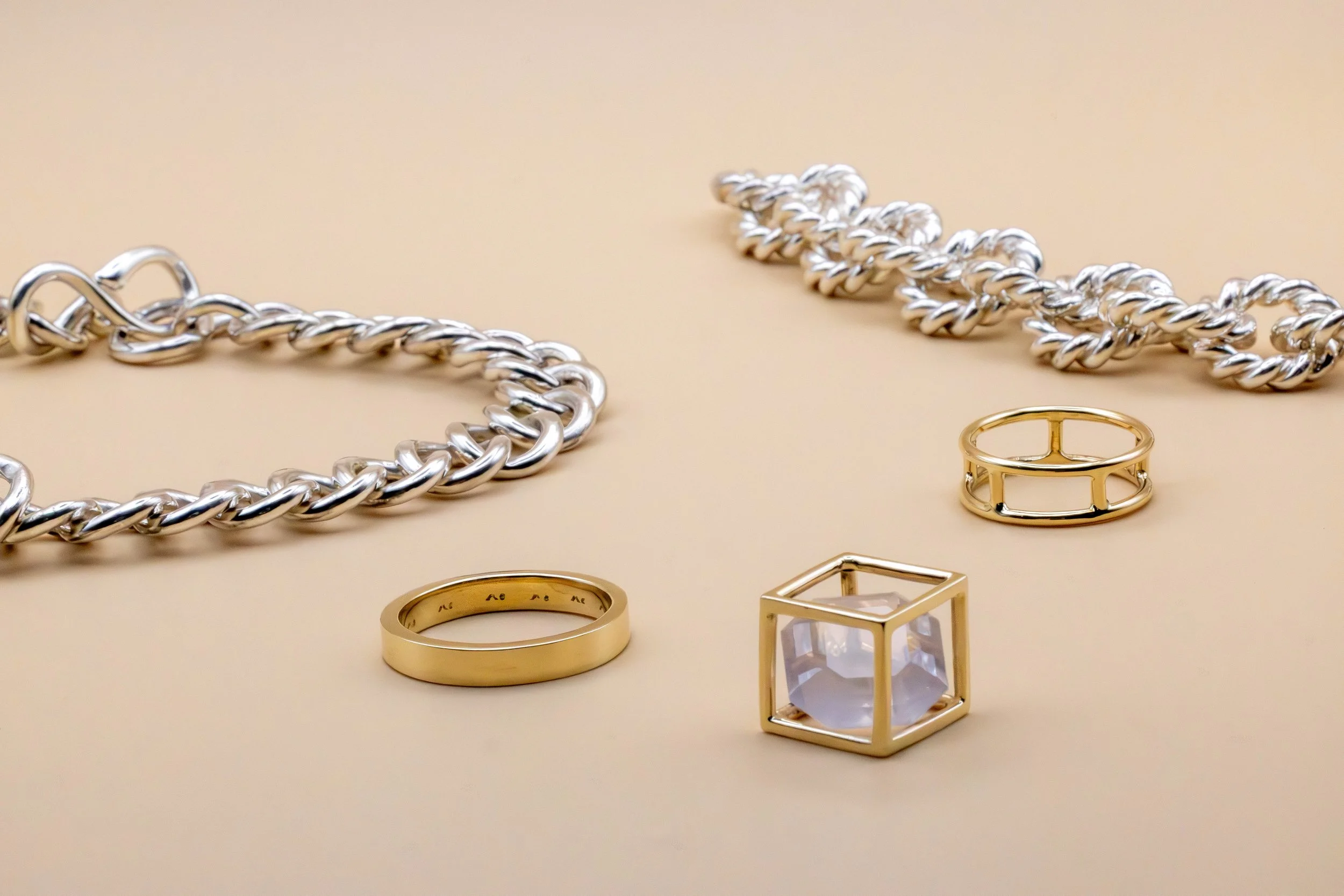 Silver and gold jewelry pieces, including silver chain necklaces, gold rings, and a golden cube on a beige background.