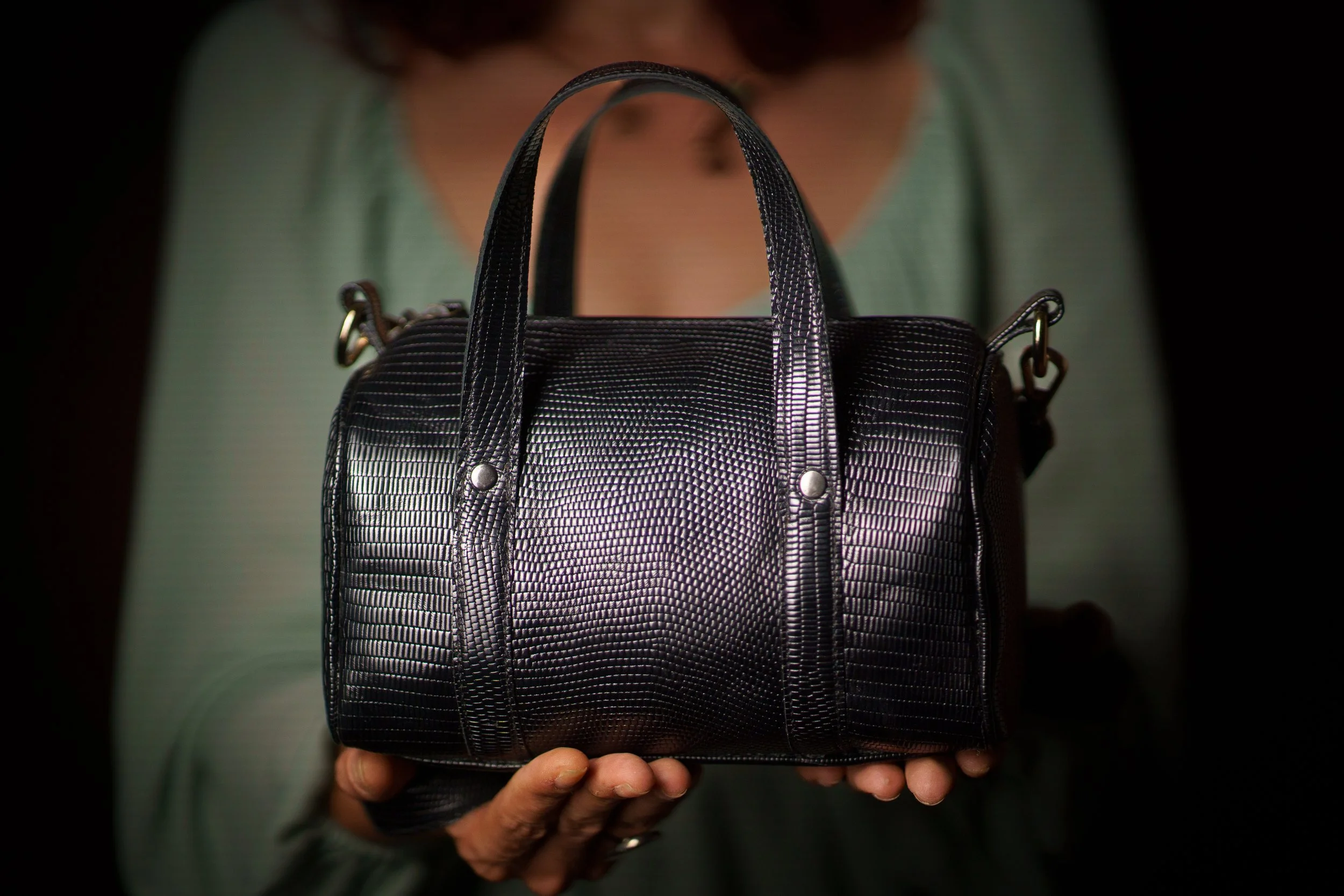 Person holding a black handbag with textured, shiny surface, metal rivets, and side hooks.