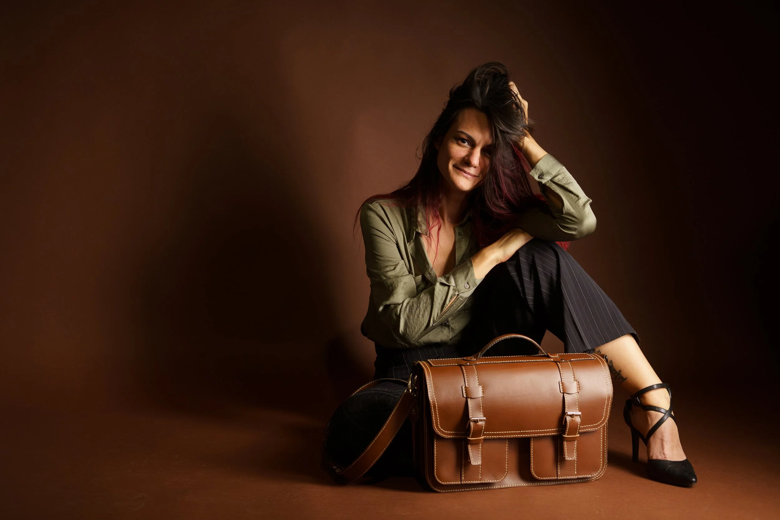A woman with dark hair and purple streaks sitting on the floor next to a leather briefcase, smiling at the camera, against a brown background.