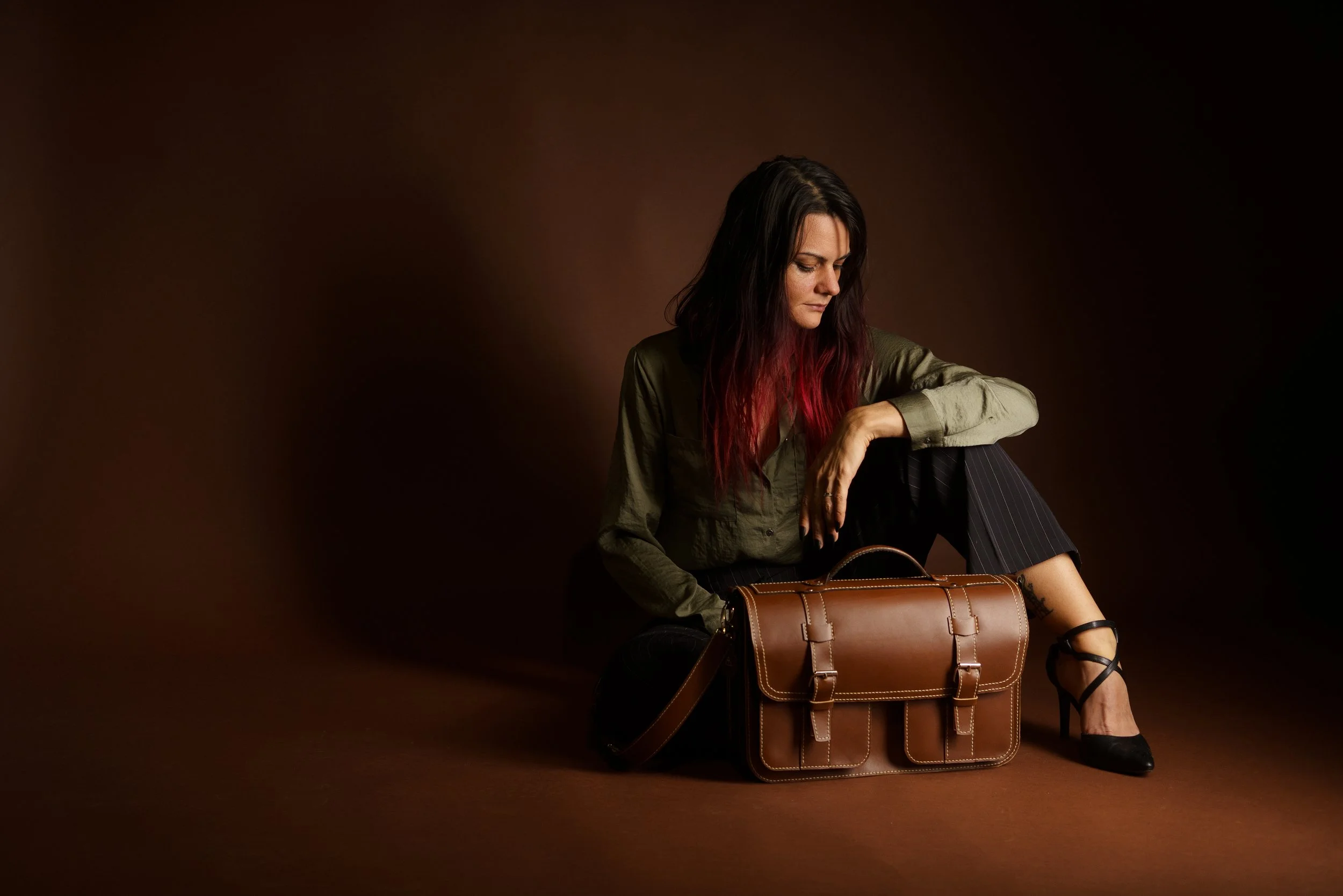 A woman with long dark hair with red tips sitting on the floor next to a brown leather bag, wearing a green shirt, black striped pants, and black high heels, in a dark studio setting.