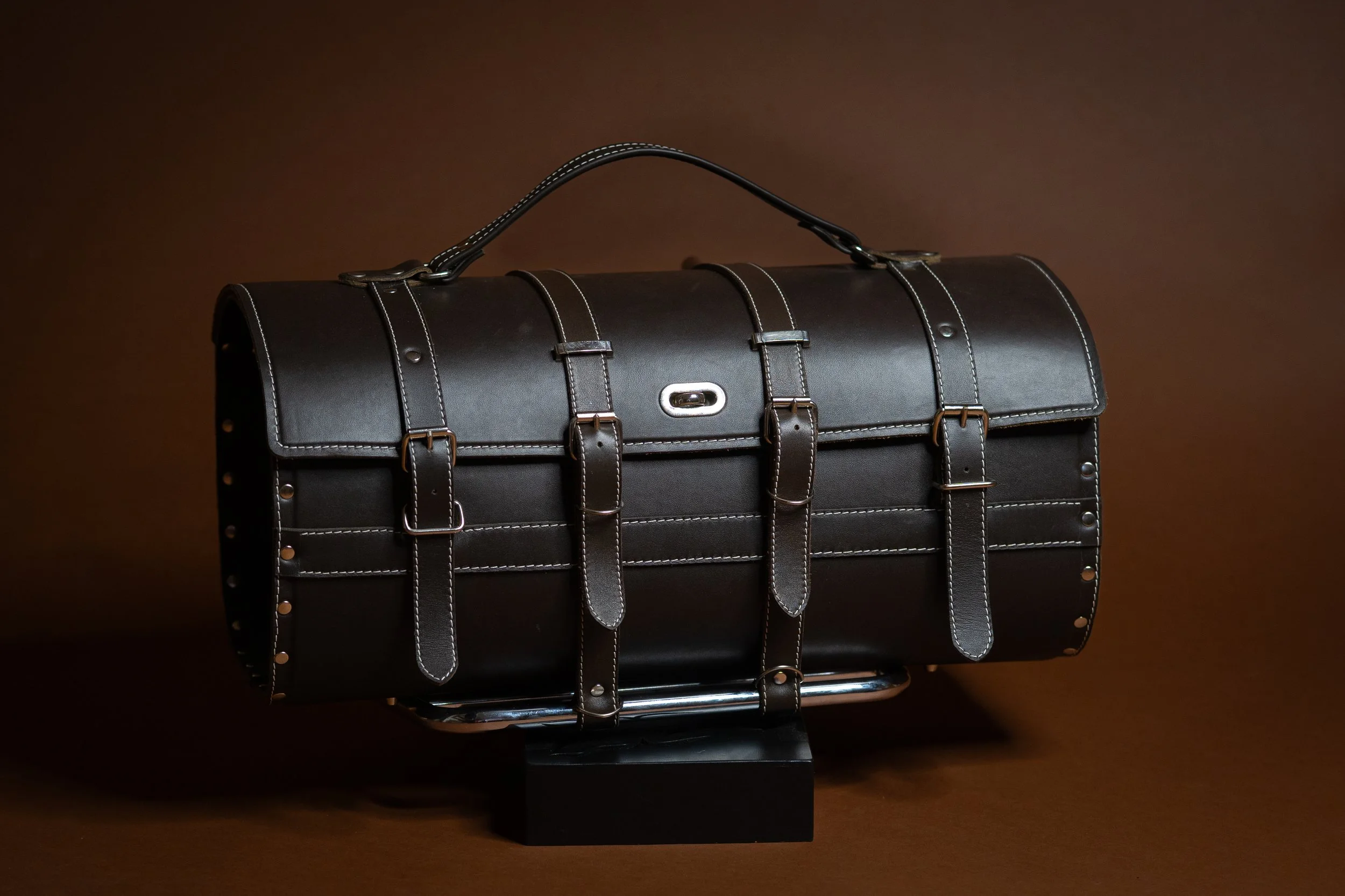 A vintage black leather doctor's bag with silver hardware and white stitching, displayed on a black stand against a brown background.