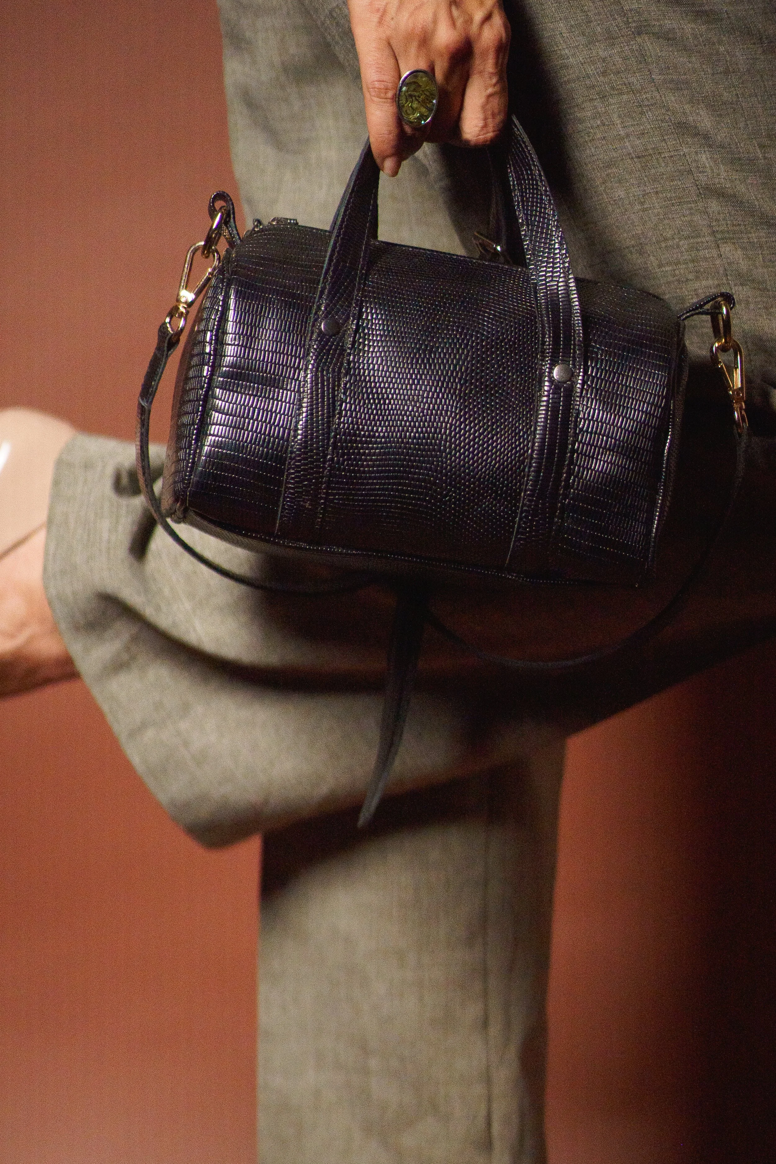 Person holding a black textured handbag with gold hardware, wearing a beige suit and a large ring with a green stone.