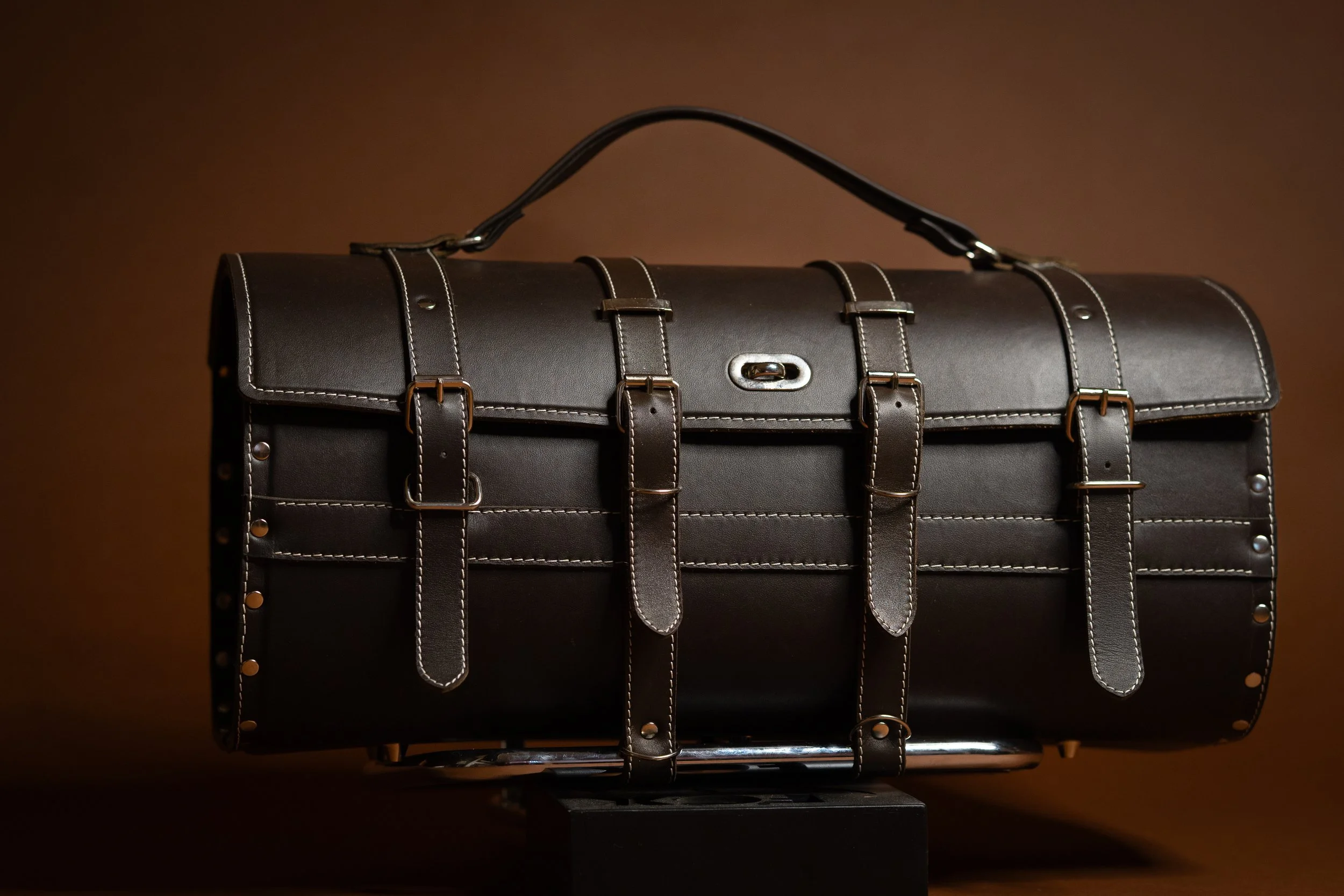 A black leather briefcase with multiple straps and metal hardware, set against a brown background.