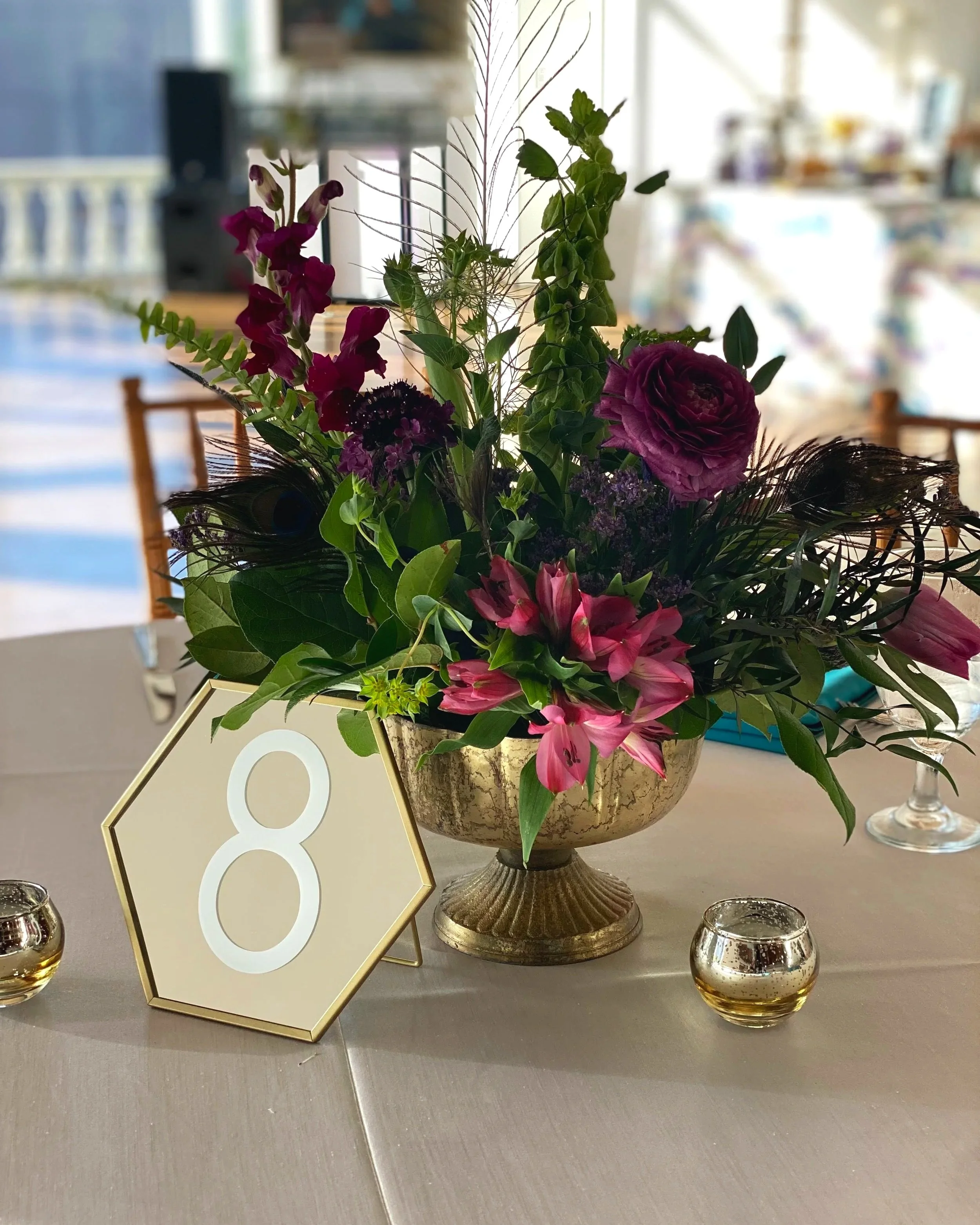 Vase with purple, pink, and magenta flowers and greenery, table number 8, and small candle holders at a formal event.